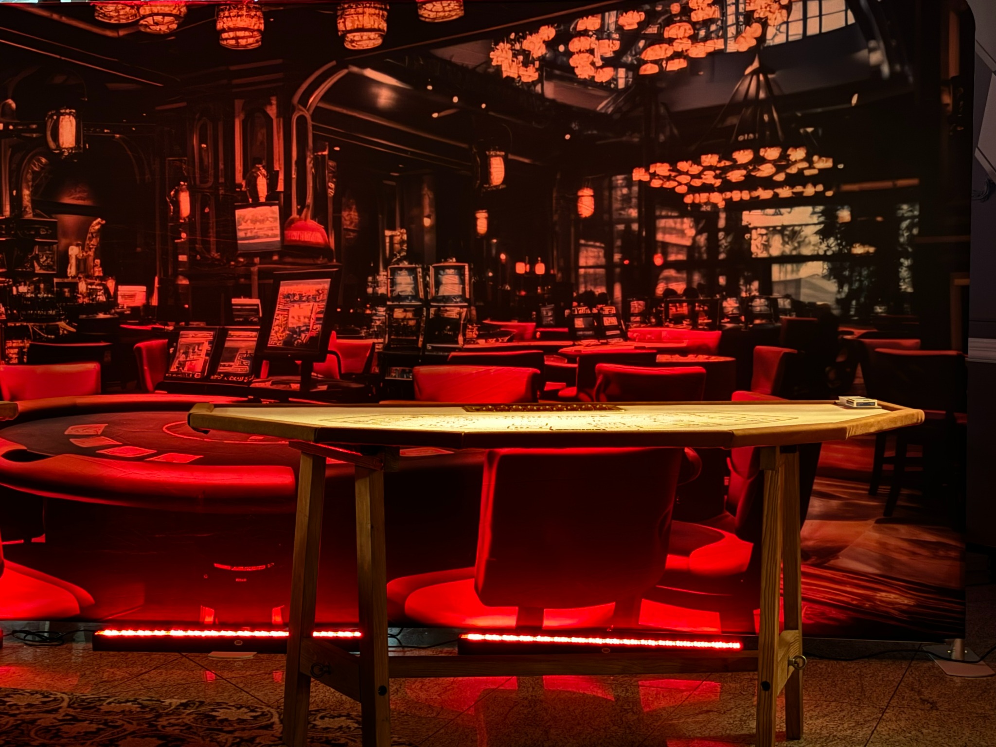 Scena z kasynem w czerwonej tonacji, stół do gry w karty oświetlony ledami, w tle dekoracja imitująca wnętrze kasyna. Klimatyczna aranżacja na imprezę tematyczną.