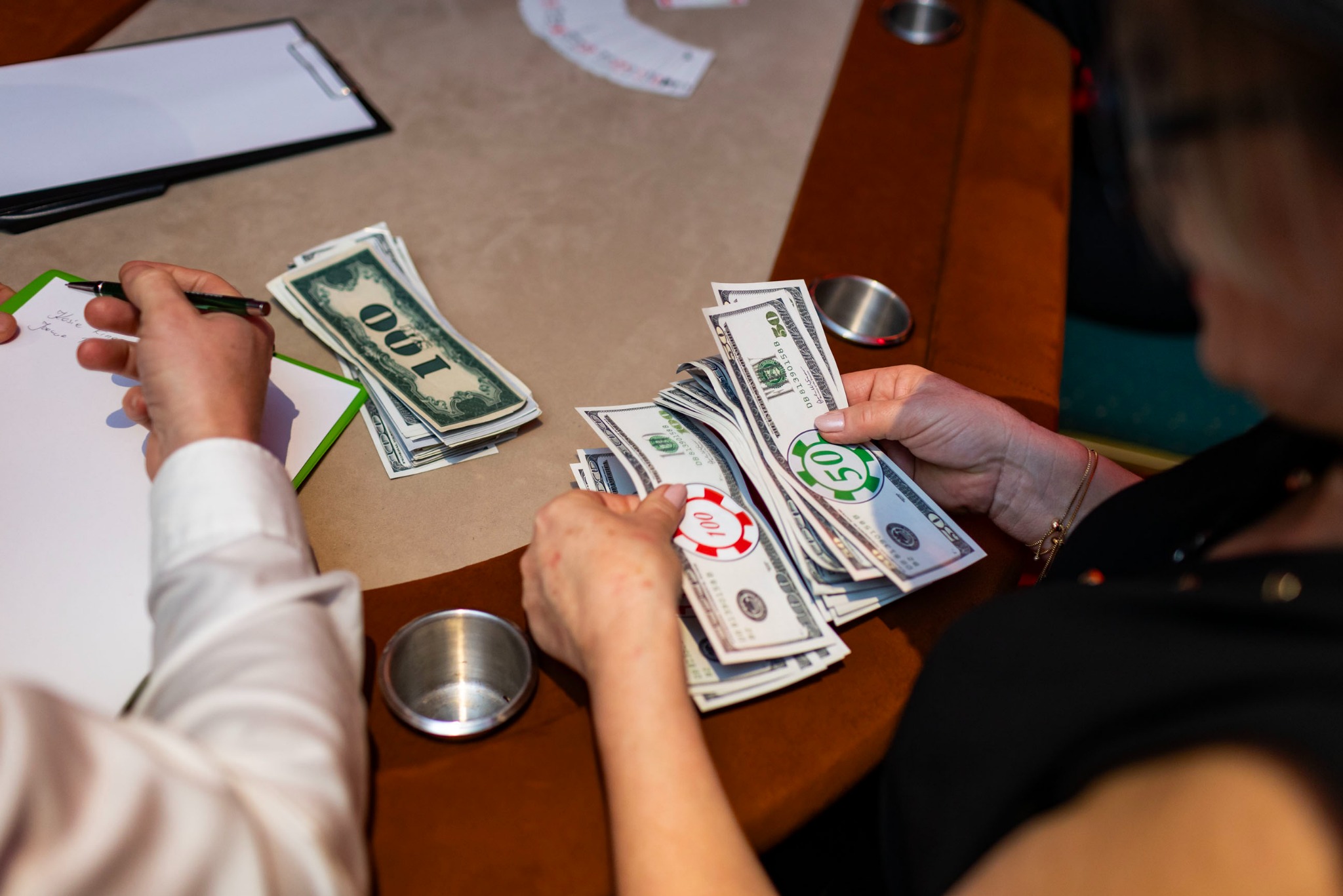 Osoby liczą pieniądze i żetony na stole do gry, prawdopodobnie podczas imprezy tematycznej w stylu kasyna. Widoczne banknoty i żetony o różnych nominałach.