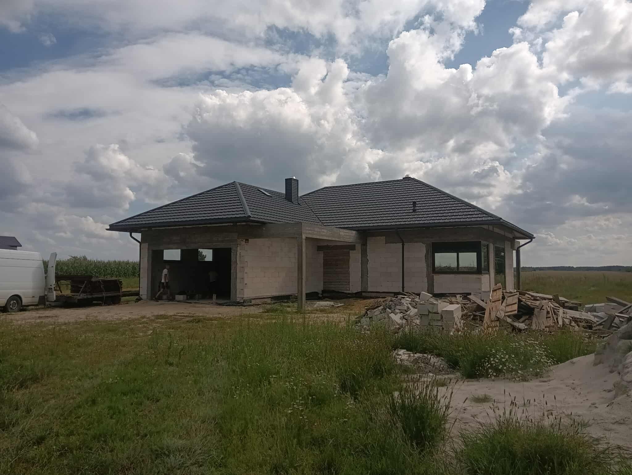 Surowy dom z szarej cegły z ciemnym dachem na tle pochmurnego nieba. Widoczne materiały budowlane i częściowo otwarty garaż. Etap budowy.