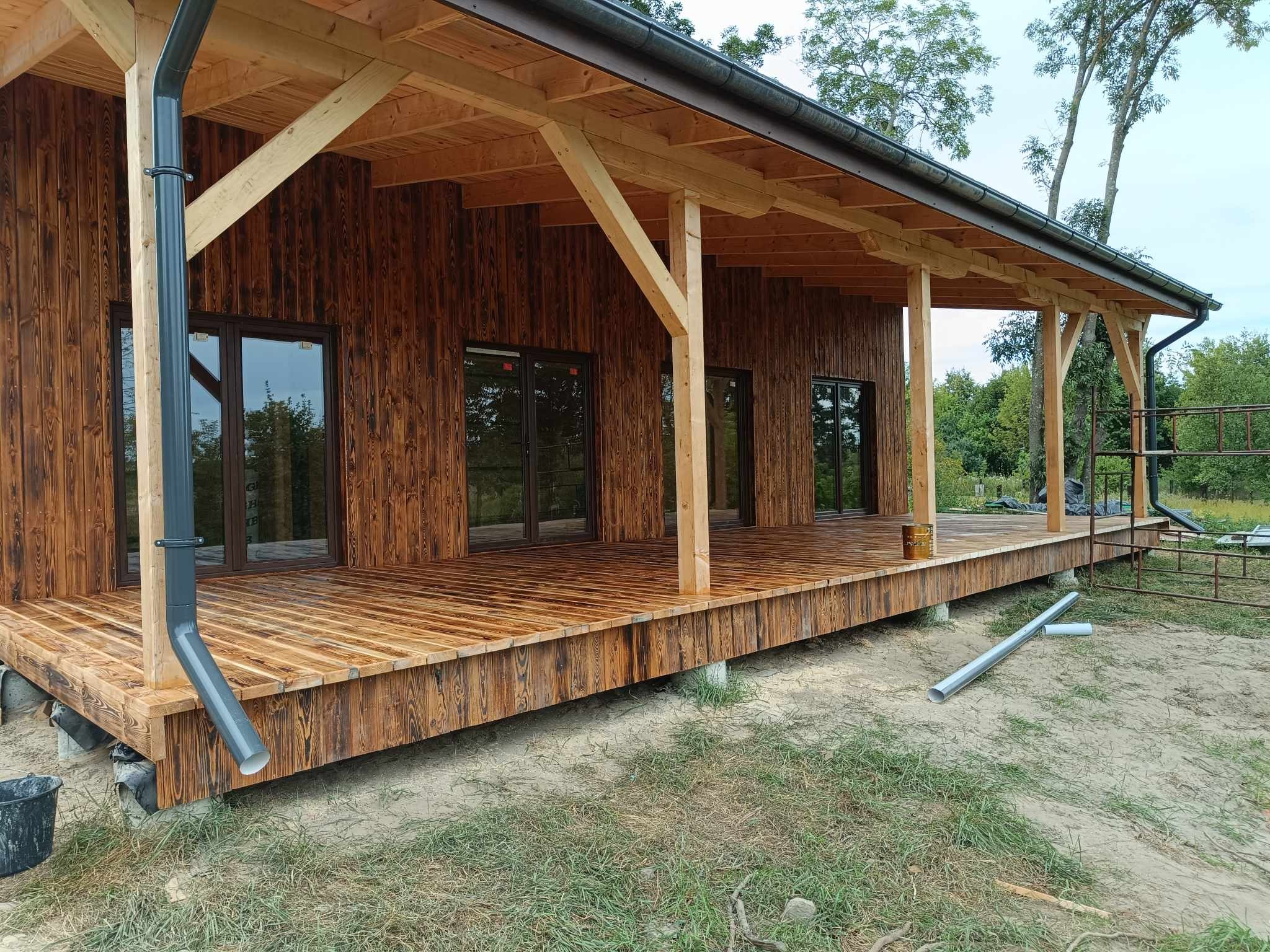 Drewniany dom z tarasem i widocznym systemem rynnowym. Świeżo zaimpregnowane drewno o ciepłym, brązowym odcieniu. Widok na zewnątrz przez okna.