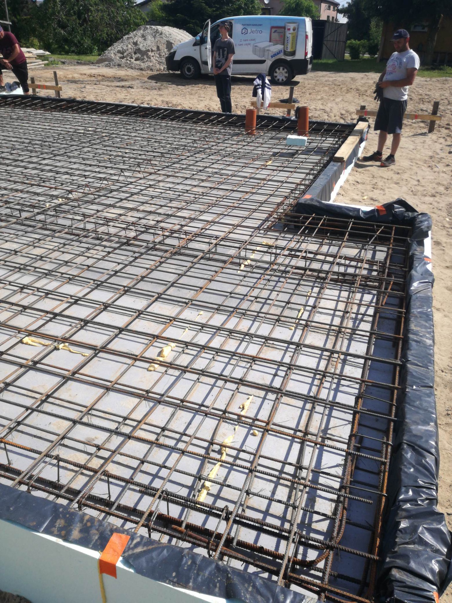 Zbrojenie fundamentu domu jednorodzinnego z widoczną izolacją termiczną i folią. W tle pracownicy i samochód dostawczy.