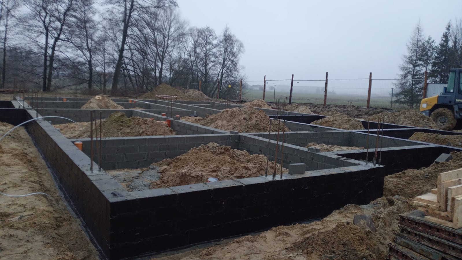 Zalane fundamenty z bloczków betonowych, zabezpieczone czarną izolacją, z widocznymi prętami zbrojeniowymi i stertami piasku wewnątrz, na tle pochmurnego krajobrazu.