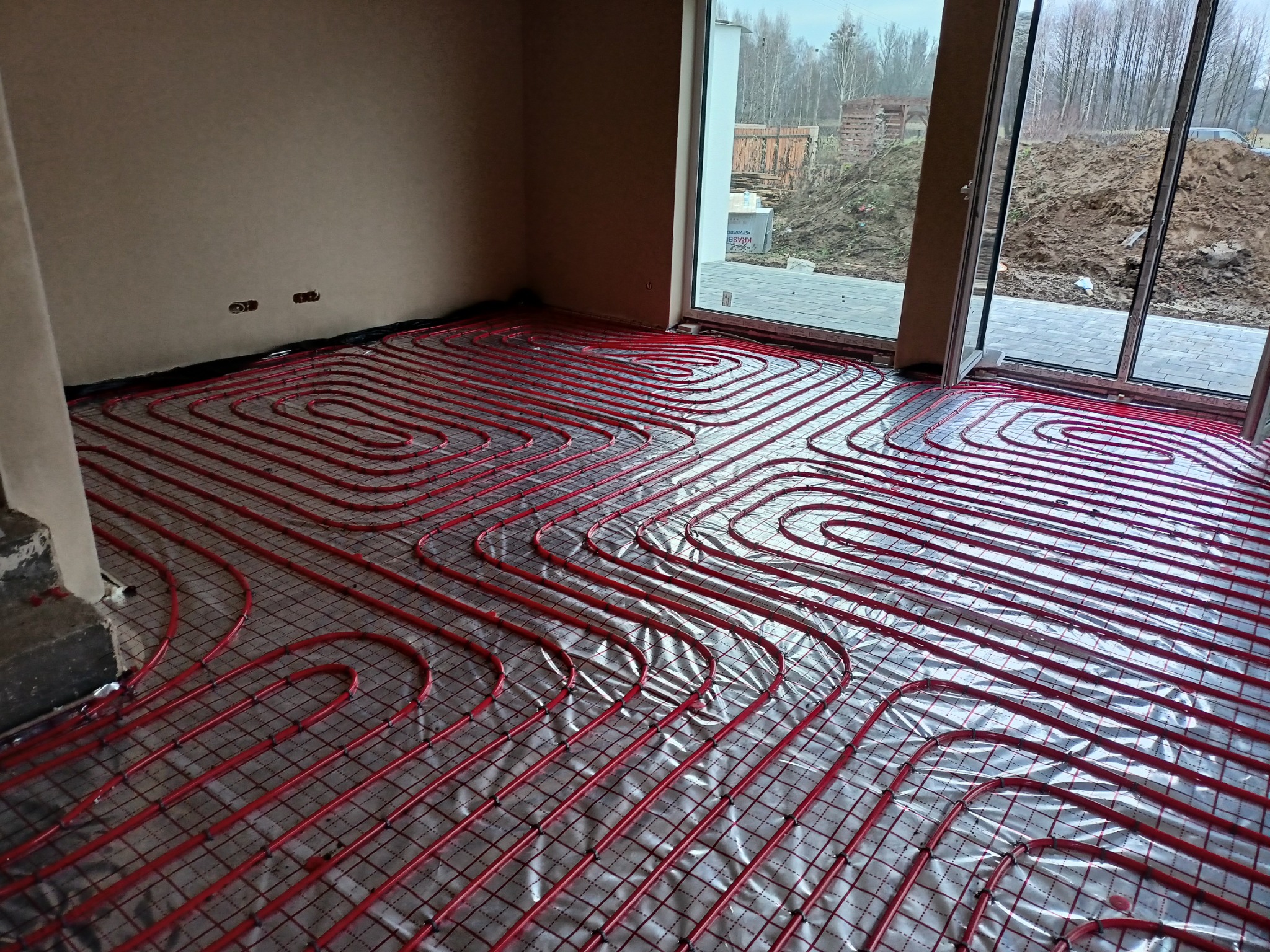 Czerwone rury ogrzewania podłogowego ułożone na folii odblaskowej w jasnym pomieszczeniu z dużym oknem, widok na plac budowy.