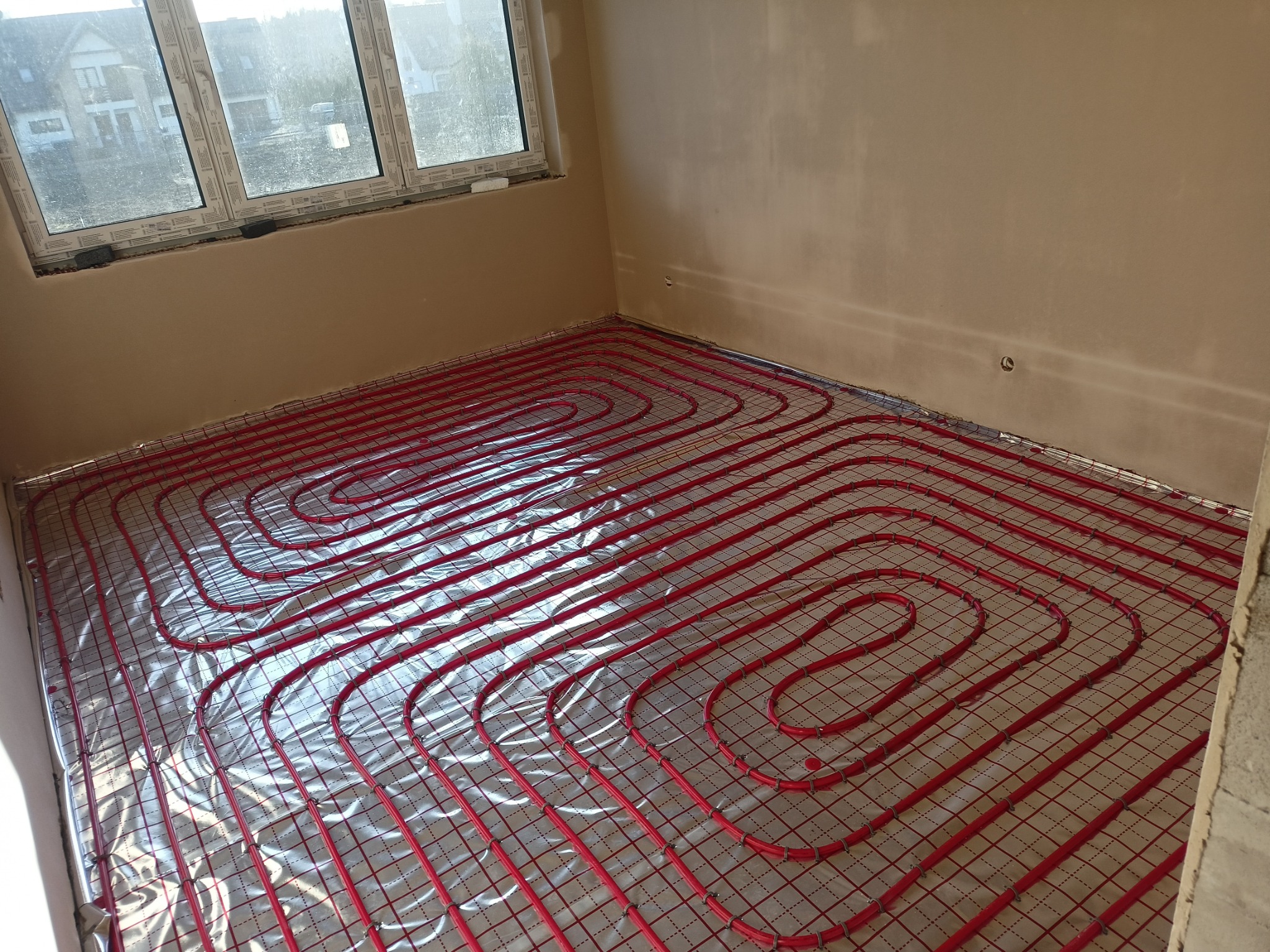Czerwone rury ogrzewania podłogowego ułożone spiralnie na srebrnej folii izolacyjnej w jasnym pomieszczeniu z oknem. Widok z góry, surowy stan wykończenia.