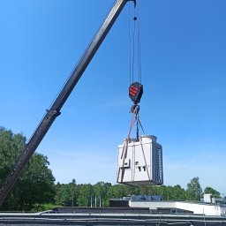 SiM Marcin Nowak - Montaż klimatyzacji na dachu budynku za pomocą dźwigu. Widok na urządzenie, dźwig i błękitne niebo. W tle zieleń drzew. Prace instalacyjne w toku.