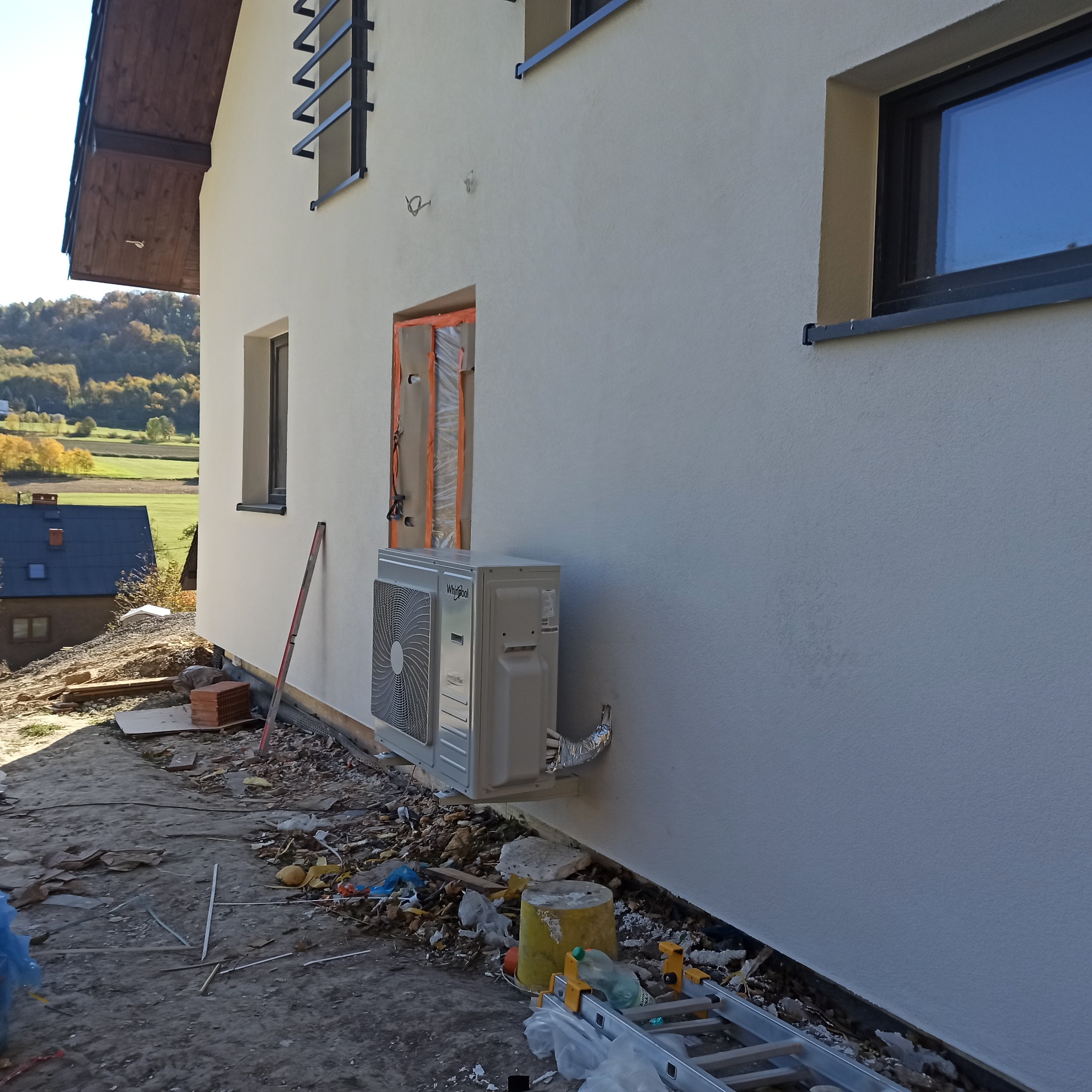 Zamontowana jednostka zewnętrzna klimatyzacji Whirlpool na elewacji domu. Widoczne przewody i elementy montażowe. W tle krajobraz i budynki.