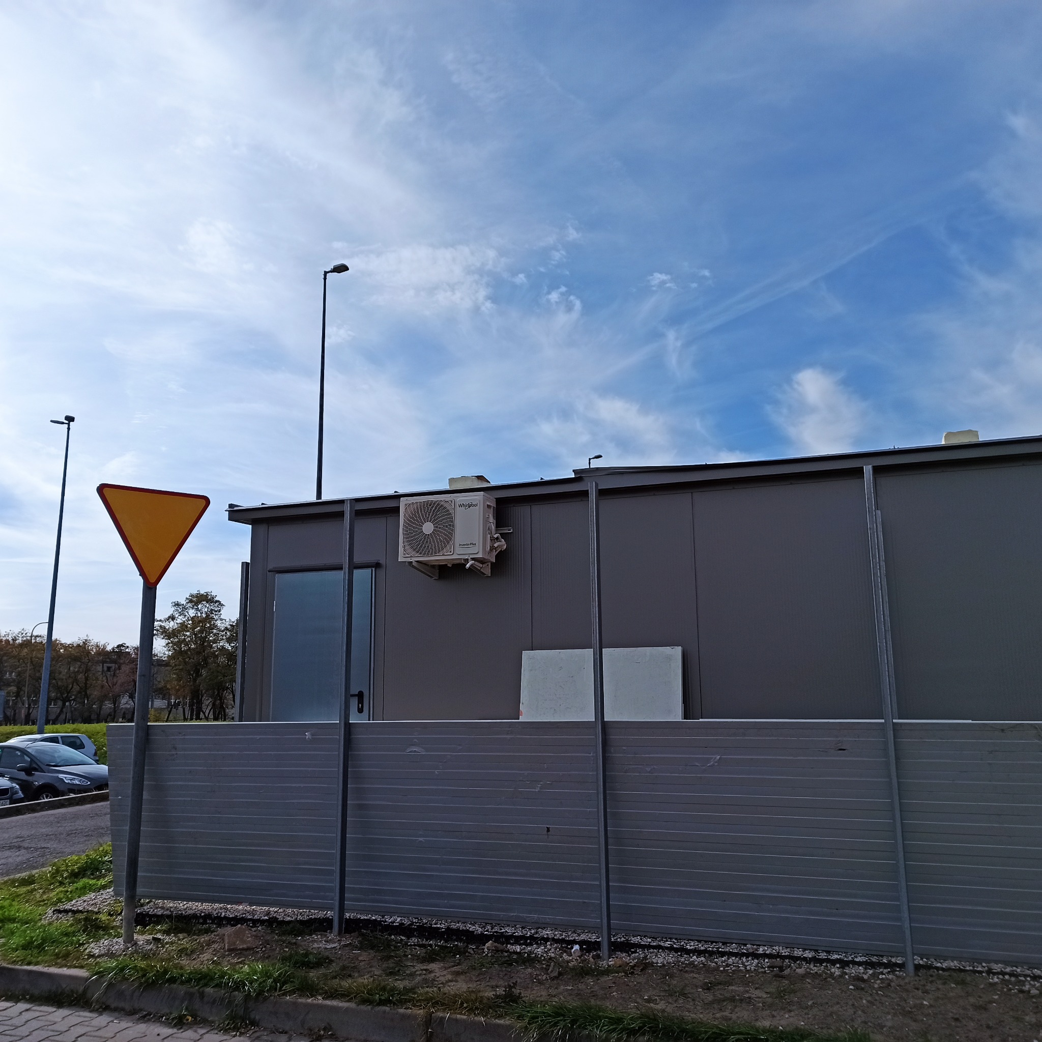 Szary kontener biurowy z zamontowaną klimatyzacją Whirlpool na ścianie, widoczny znak drogowy i błękitne niebo z chmurami w tle.