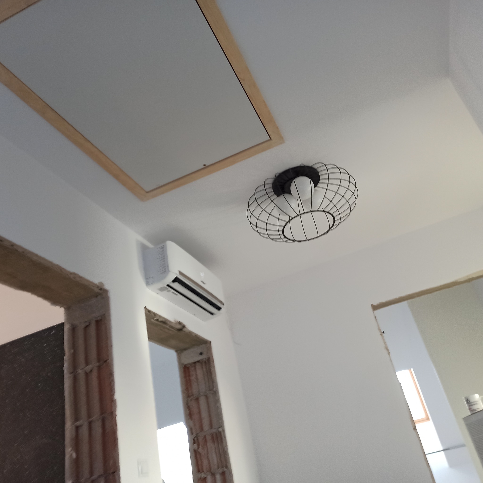 Biała klimatyzacja na ścianie w trakcie remontu, widoczna lampa i sufit z klapą rewizyjną. Ujęcie z dołu, podkreślające etap prac wykończeniowych.