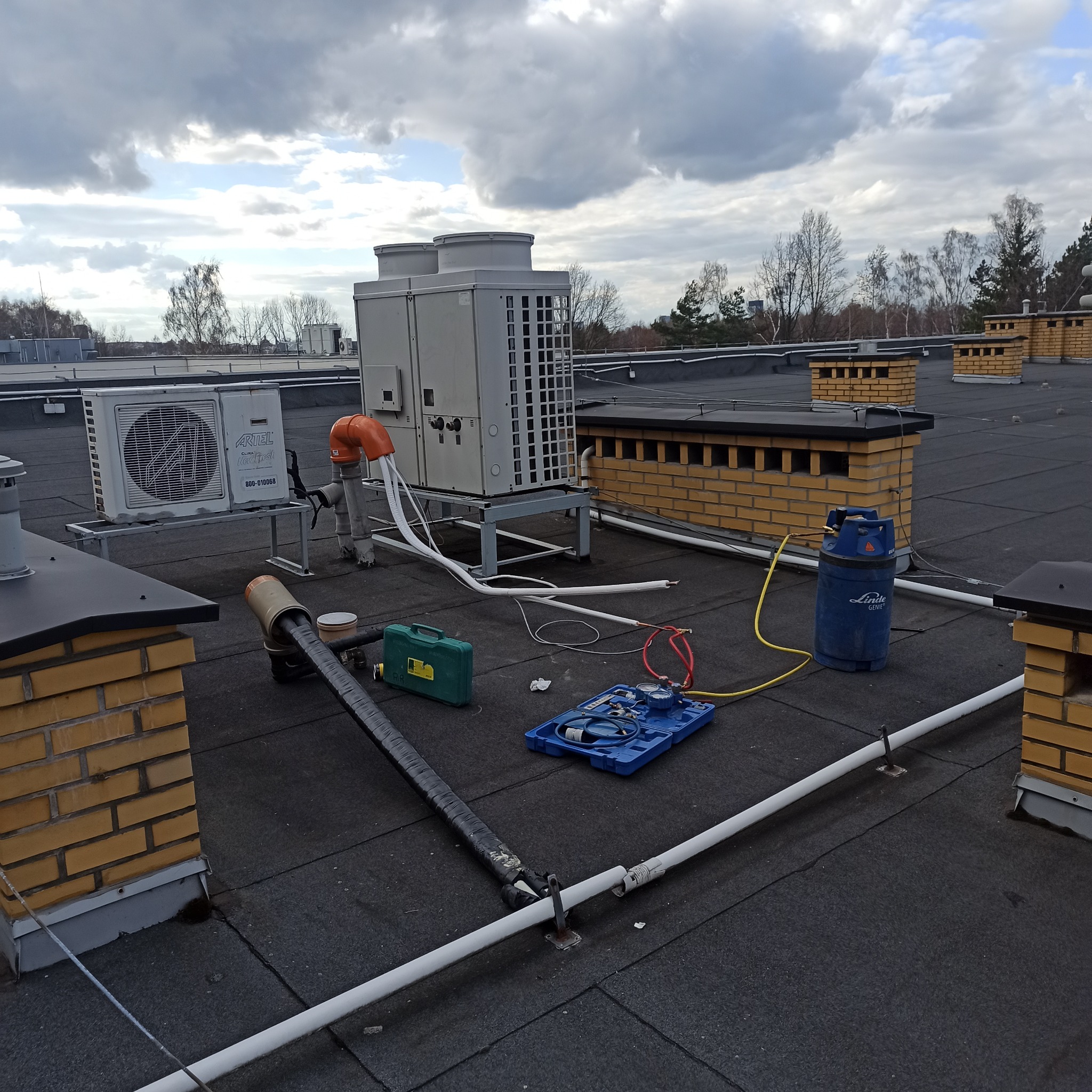 Klimatyzacja na dachu budynku, widoczne narzędzia serwisowe i butla z gazem. Prace serwisowe w toku, pochmurne niebo w tle.