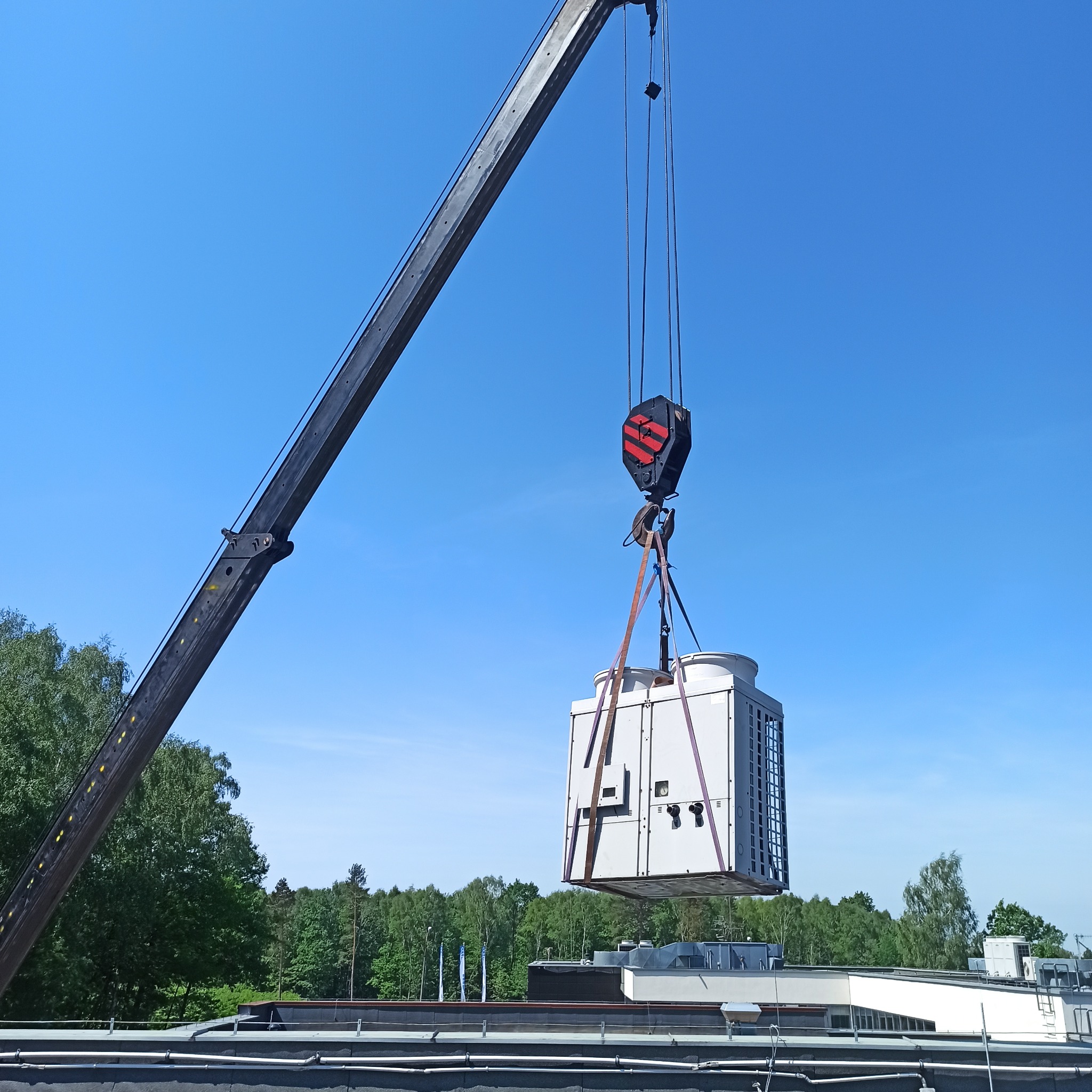 Montaż klimatyzacji na dachu budynku za pomocą dźwigu. Widok na urządzenie, dźwig i błękitne niebo. W tle zieleń drzew. Prace instalacyjne w toku.
