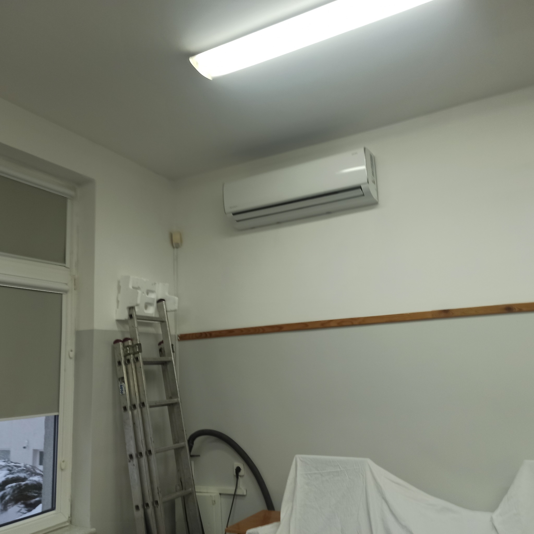 Biały klimatyzator ścienny w jasnym pomieszczeniu, widoczna drabina i zabezpieczone meble, sugerujące montaż lub serwis.