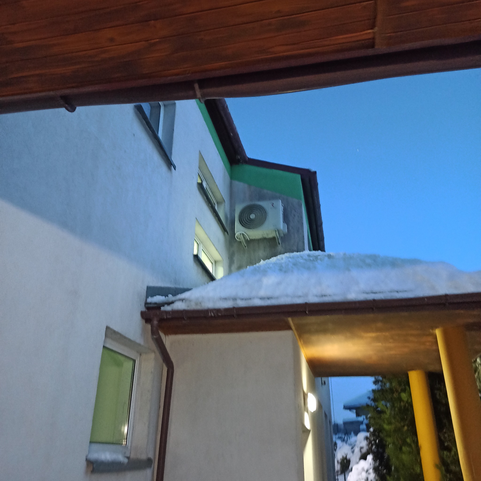 Biała jednostka klimatyzacyjna zamontowana na ścianie budynku, częściowo zasłonięta śniegiem na dachu. Widok z dołu, niebieskie niebo w tle.