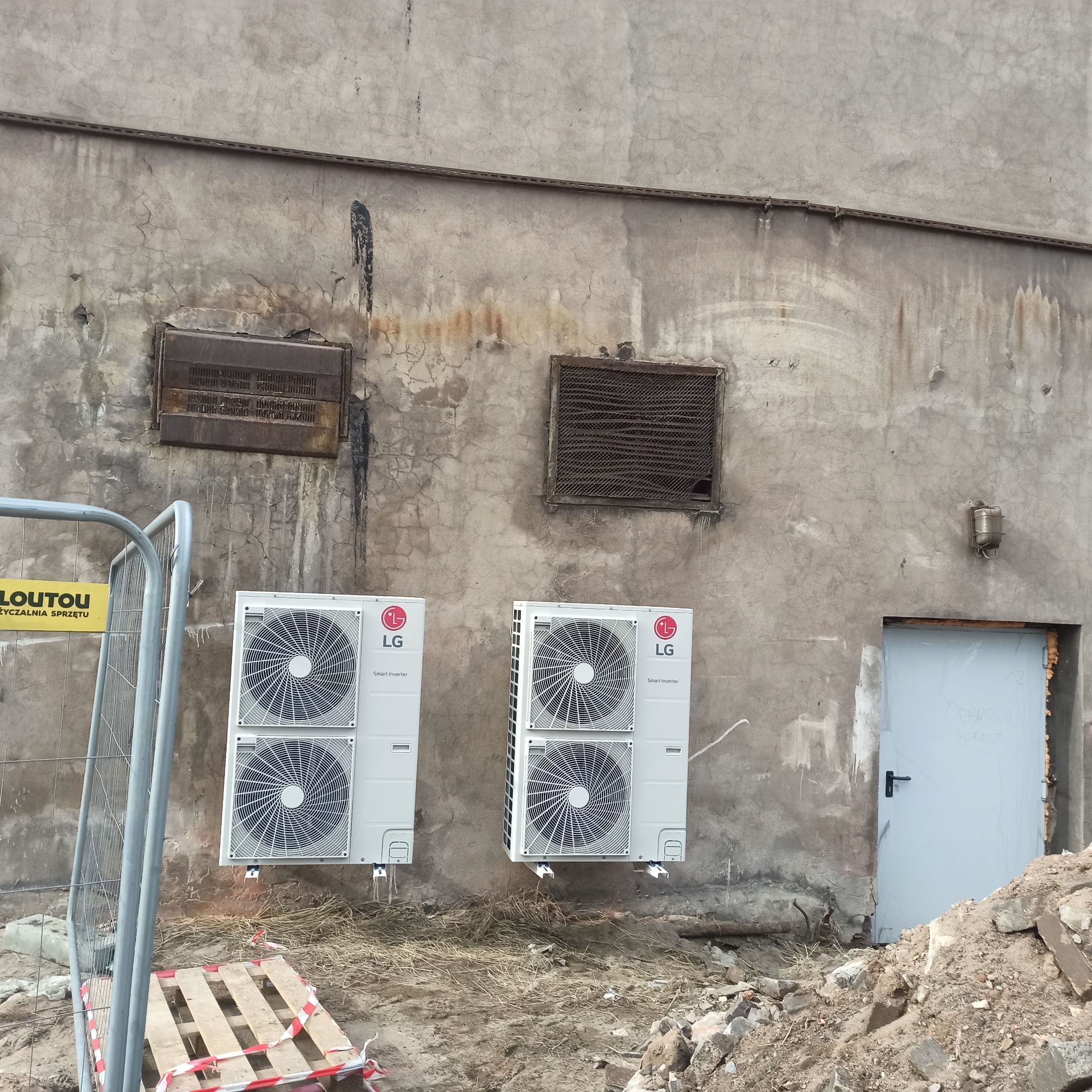 Dwa białe klimatyzatory LG zamontowane na szarej, zniszczonej ścianie budynku. Widoczne stare kratki wentylacyjne i gruz na ziemi.