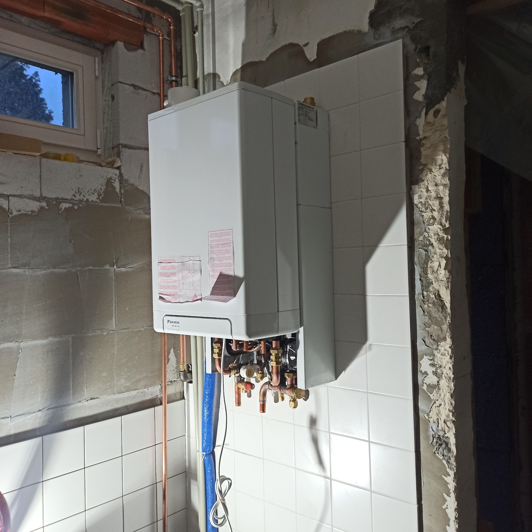 Biały, wiszący piec gazowy Daikin Altherma Hybrid Heat Pump z widocznymi miedzianymi rurami instalacji, zamontowany na ścianie z białych płytek w surowym wnętrzu.