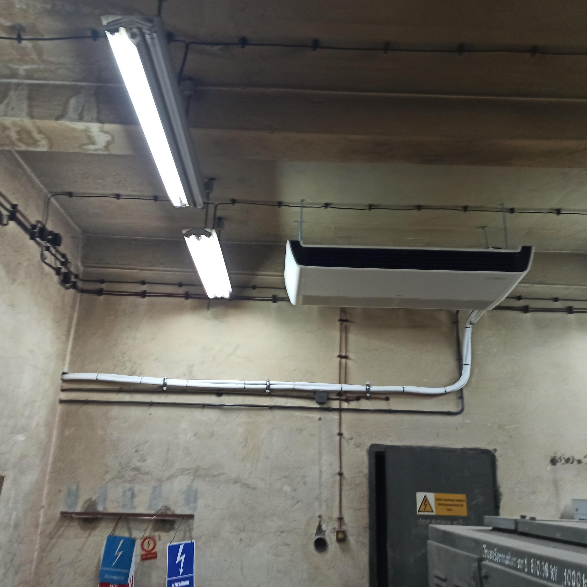 Zamontowana klimatyzacja kasetonowa w pomieszczeniu z widocznymi instalacjami elektrycznymi i rurami. Surowy, industrialny charakter wnętrza.