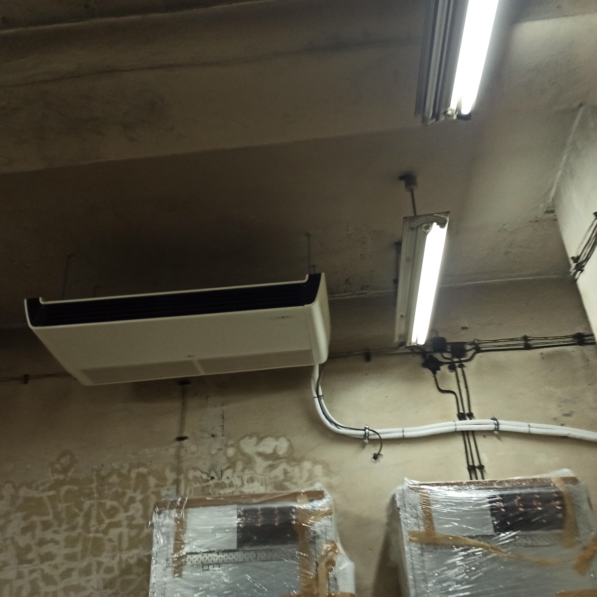 Zamontowana klimatyzacja kasetonowa pod sufitem, widoczne przewody i lampa. Na dole dwa urządzenia w folii. Ściana z widocznymi śladami wilgoci.