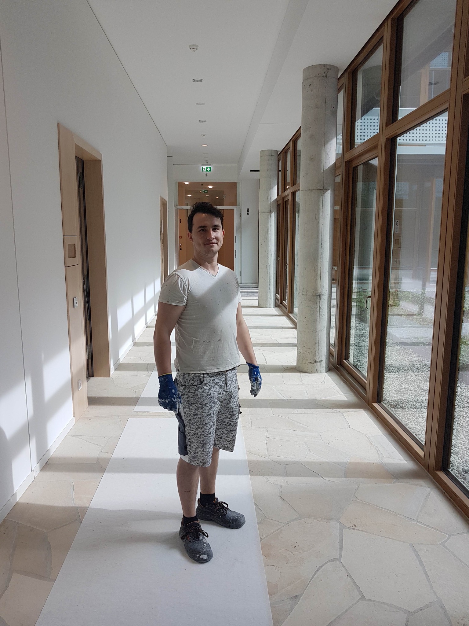 Mężczyzna w ubrudzonym farbą t-shircie i rękawicach malarskich stoi w korytarzu z pomalowaną podłogą, na tle białych ścian i okien z drewnianą ramą.