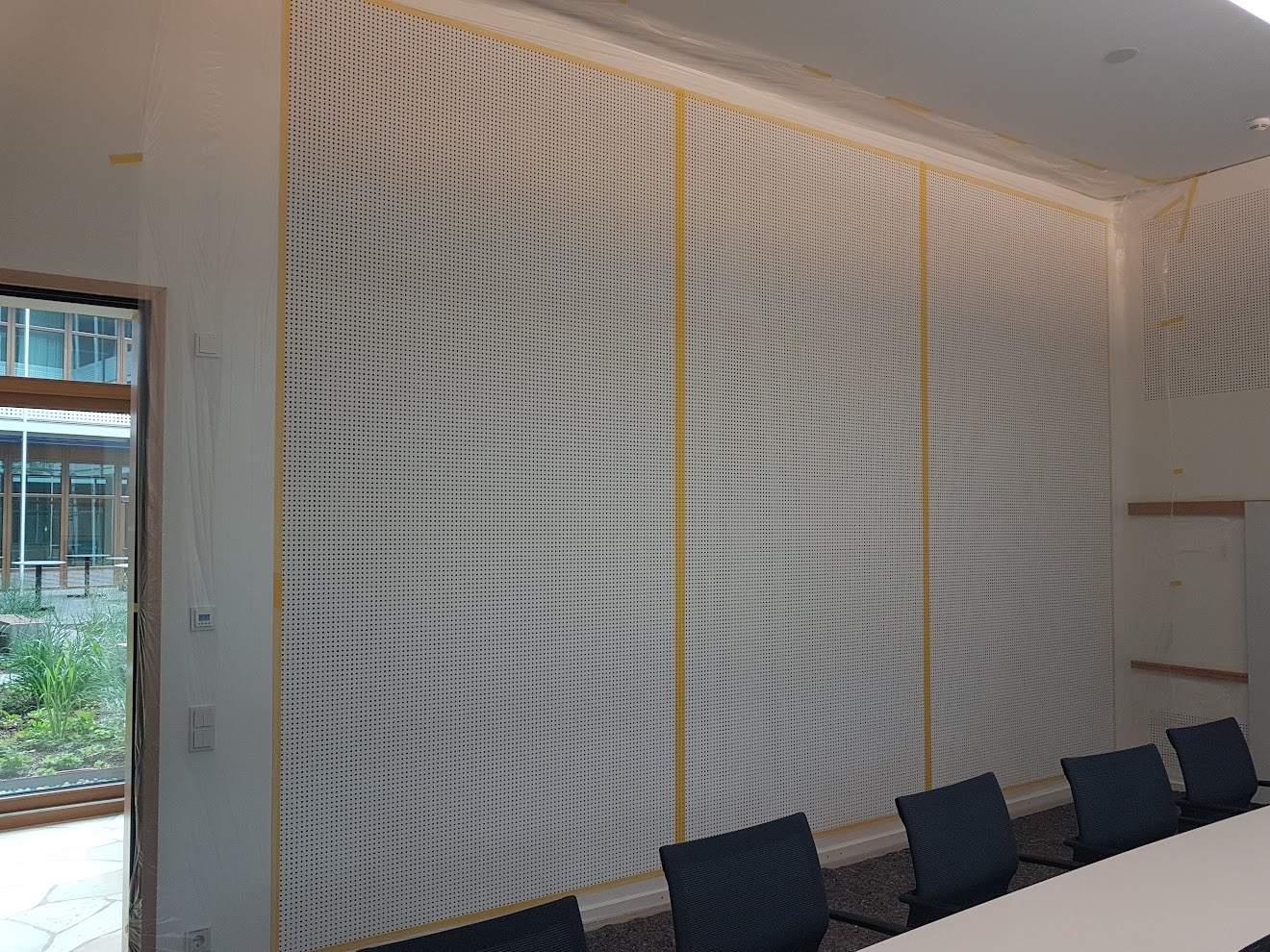 Biała ściana z perforacją zabezpieczona folią i taśmą malarską przed malowaniem w biurze. Widoczne krzesła i stół konferencyjny.