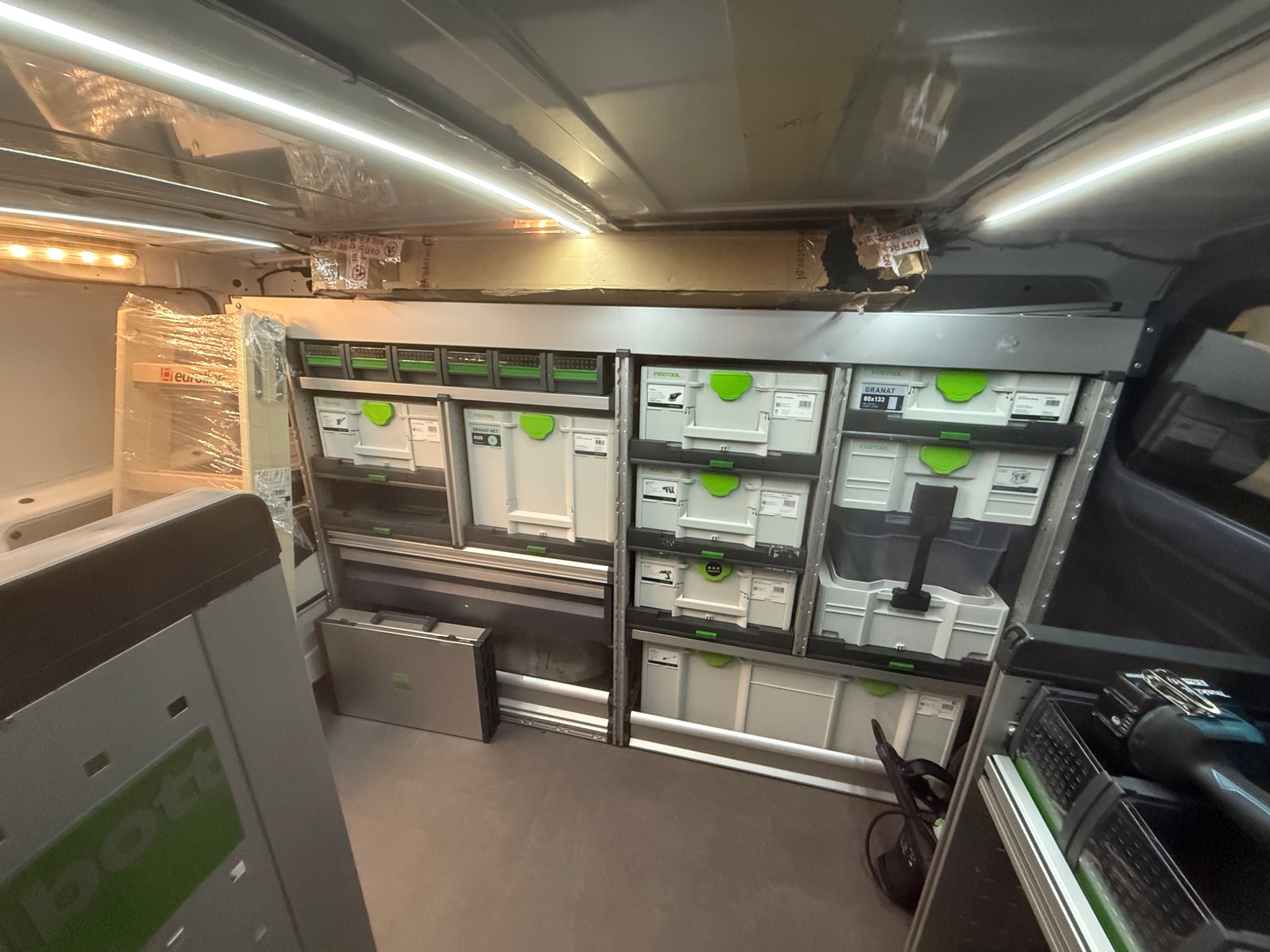 Wnętrze furgonetki z zabudową warsztatową: regały z szufladami i skrzynkami narzędziowymi Festool. Widoczne narzędzia i oświetlenie LED. Profesjonalne wyposażenie mobilnego warsztatu.