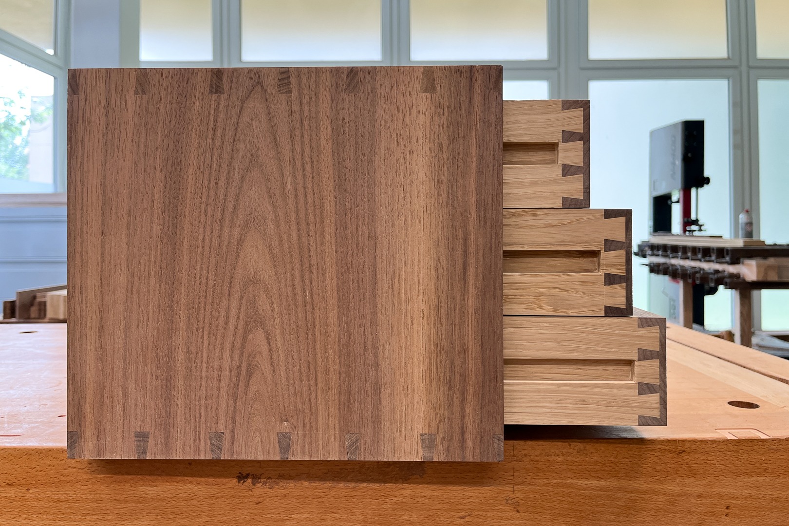 Drewniana komoda z widocznymi, precyzyjnymi połączeniami na jaskółczy ogon, wykonana z jasnego i ciemnego drewna, na tle warsztatu.