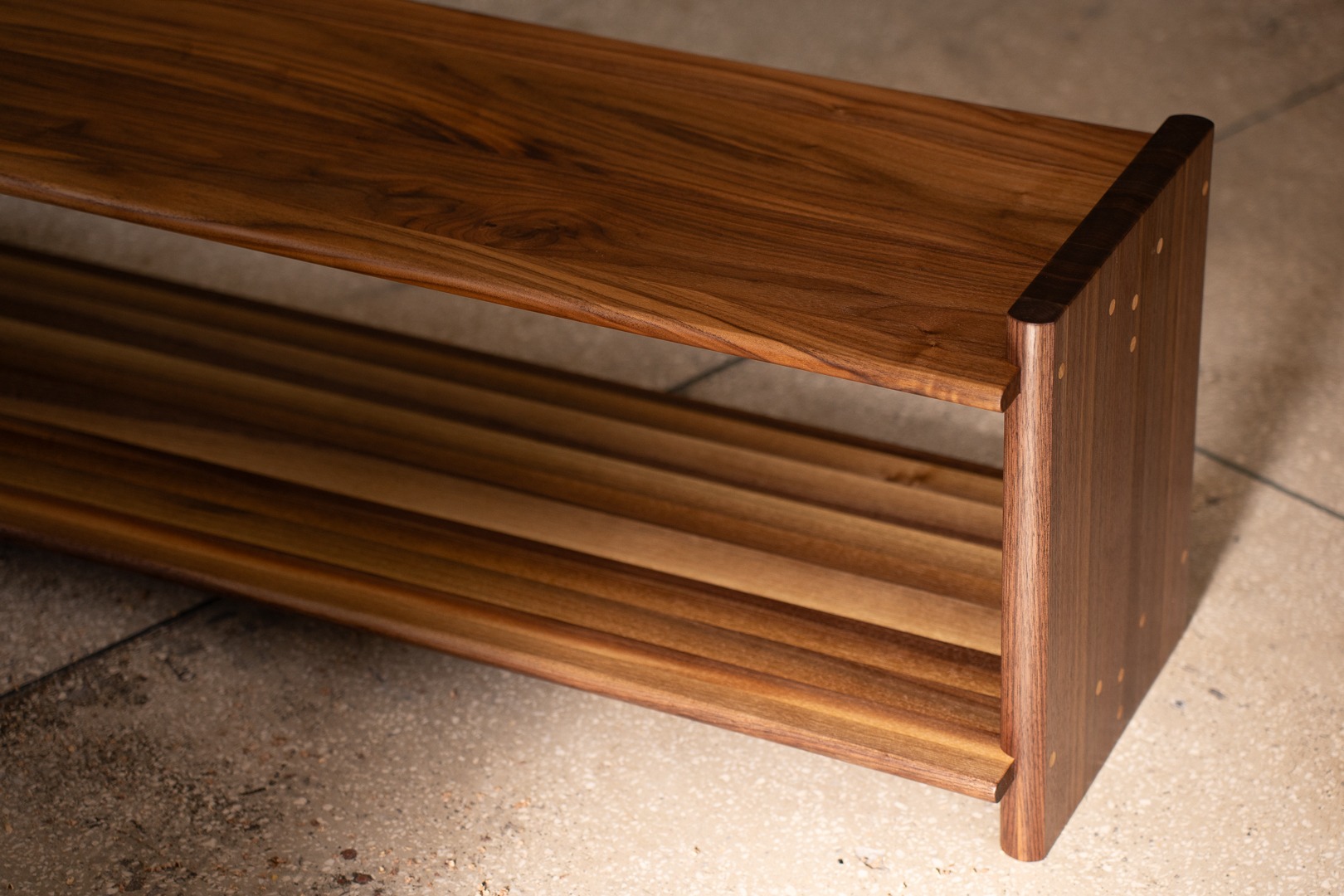 Ręcznie wykonana, drewniana półka na buty z orzecha włoskiego, z widocznymi detalami łączeń na bokach, na tle betonowej posadzki. Minimalistyczny design.
