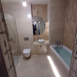 HQ Bartosz Pilarczyk - Jasna łazienka po remoncie z kabiną prysznicową, toaletą, okrągłym lustrem i wanną z podświetleniem LED. Ściany wyłożone płytkami w odcieniach beżu z wzorem jodełki.