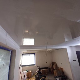 HQ Bartosz Pilarczyk - Świeżo nałożona, błyszcząca gładź na suficie w remontowanym pokoju. Widoczne okna, sprzęt malarski i przygotowane ściany. Jasne wnętrze w trakcie prac wykończeniowych.