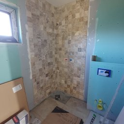 HQ Bartosz Pilarczyk - Fragment łazienki w trakcie wykańczania: ułożone płytki mozaikowe na ścianie prysznica, obok ściana z płyty gipsowej z instalacją podtynkową WC. Widoczne kartony z płytkami.