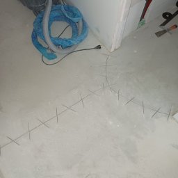 HQ Bartosz Pilarczyk - Przygotowanie podłoża pod wylewkę: zaznaczone krzyżykami punkty poziomowania, odkurzacz przemysłowy i narzędzia glazurnicze w tle. Wnętrze w trakcie prac.