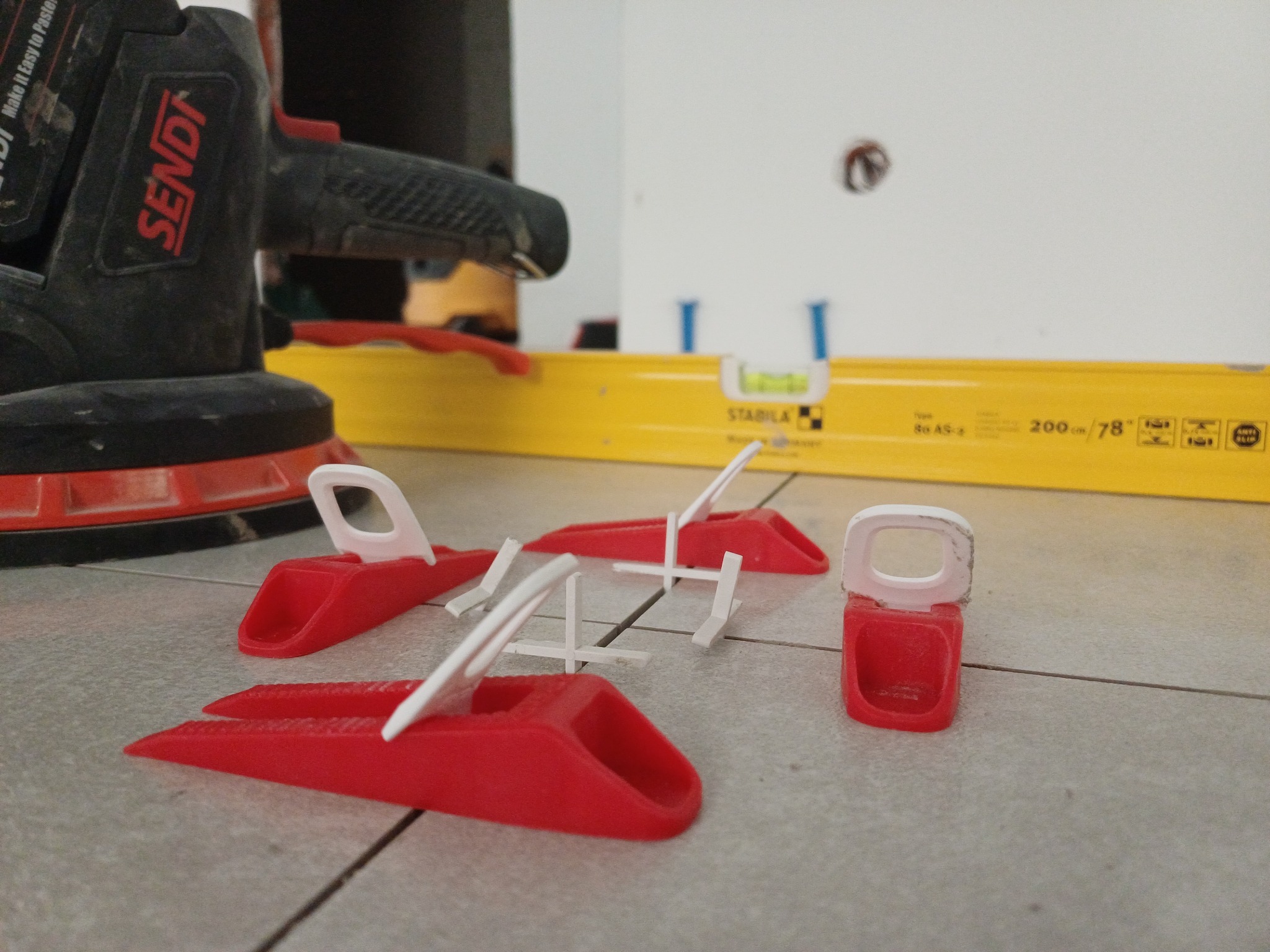 Niwelatory do płytek, poziomica i szlifierka na szarych płytkach. Narzędzia glazurnicze w zbliżeniu, widoczne detale i faktura płytek.