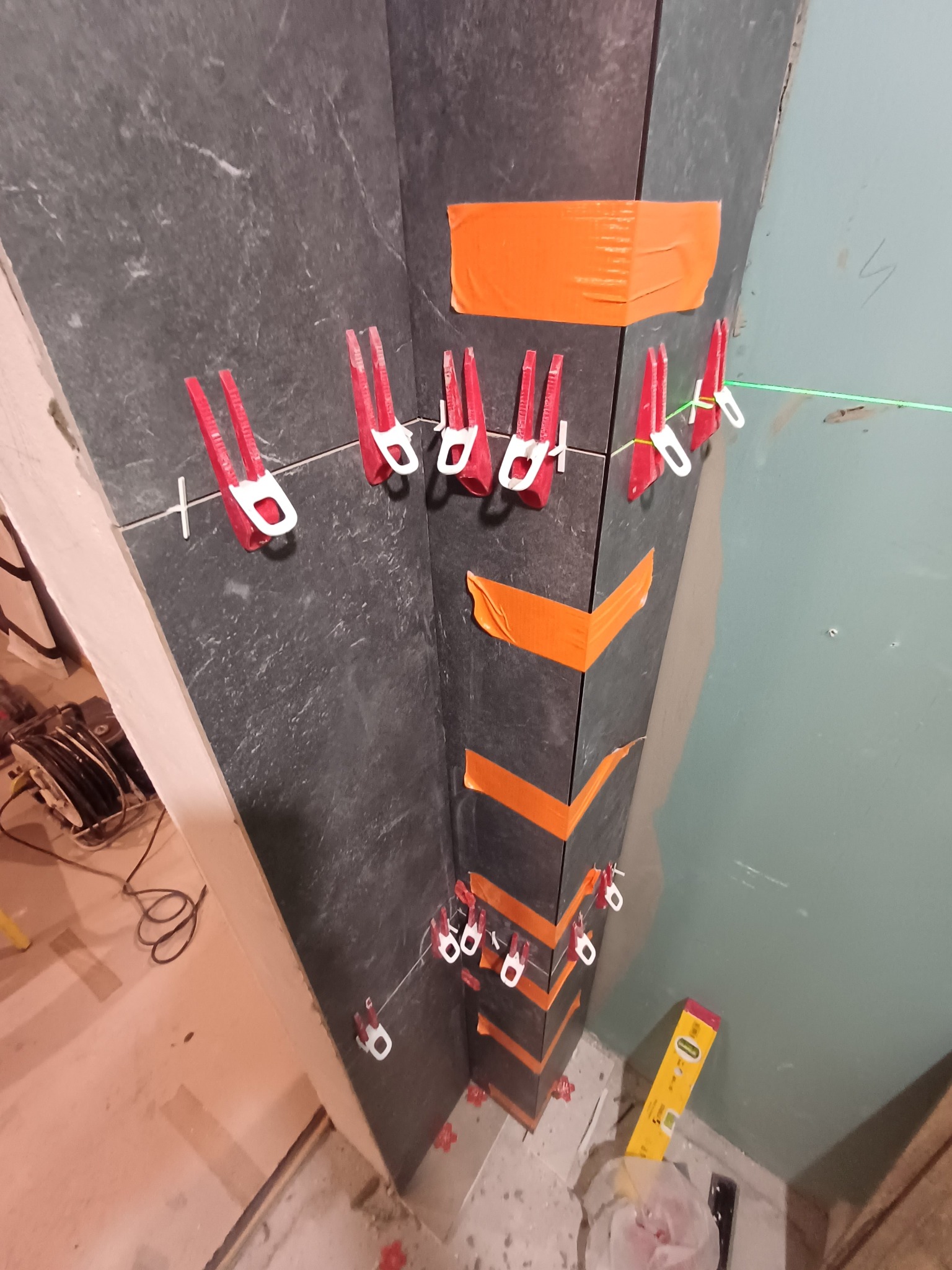 Układanie ciemnych płytek w rogu pomieszczenia, z użyciem klipsów poziomujących i pomarańczowej taśmy. Widoczny laser krzyżowy i poziomica.