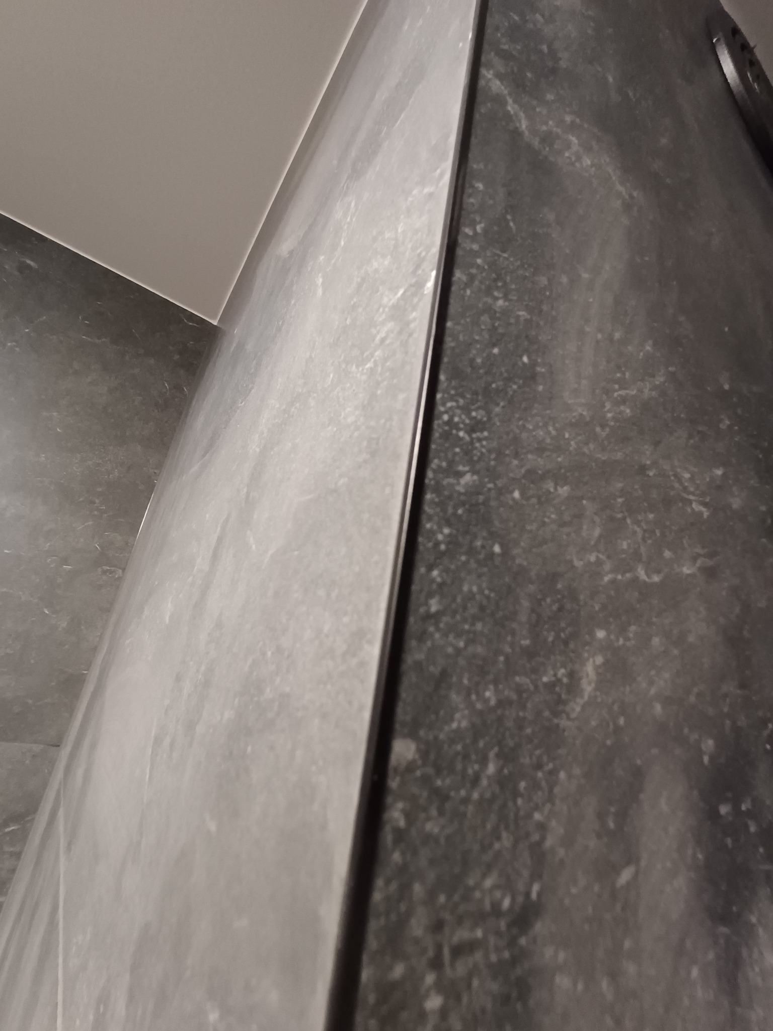 Wykończona ściana łazienki z szarych, marmurkowych płytek, widoczna pod kątem, z delikatnym oświetleniem i fugą. Minimalistyczny design.