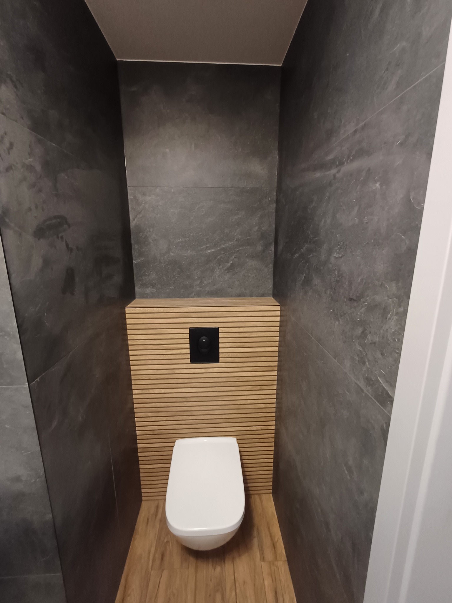 Nowoczesna toaleta wnękowa z szarą okładziną ścienną imitującą kamień i drewnianą zabudową stelaża, na podłodze panele imitujące drewno. Minimalistyczny design.