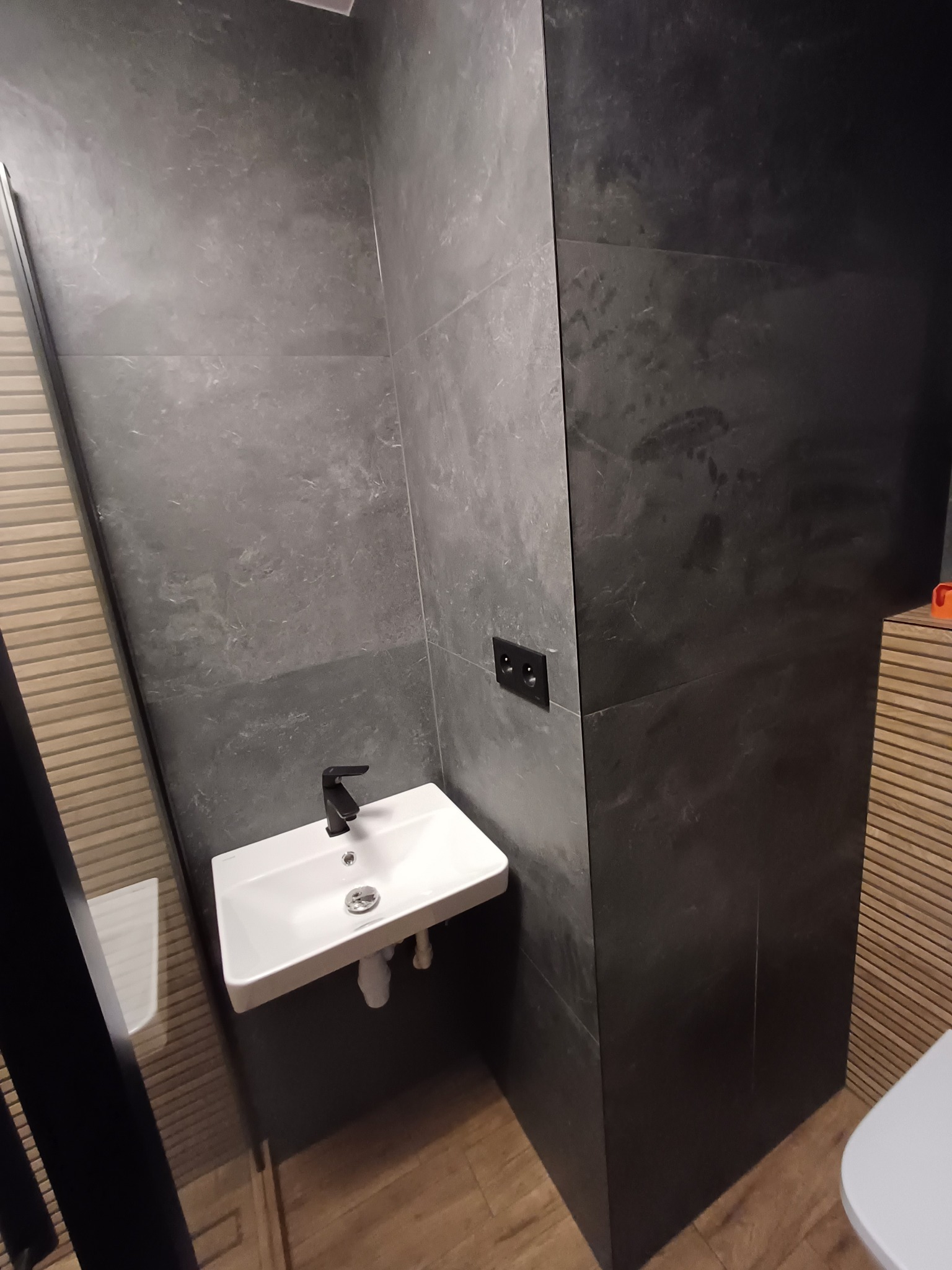 Nowoczesna łazienka z umywalką nablatową i czarną armaturą na tle ciemnych, marmurkowych płytek. Widoczne drewniane panele dekoracyjne.