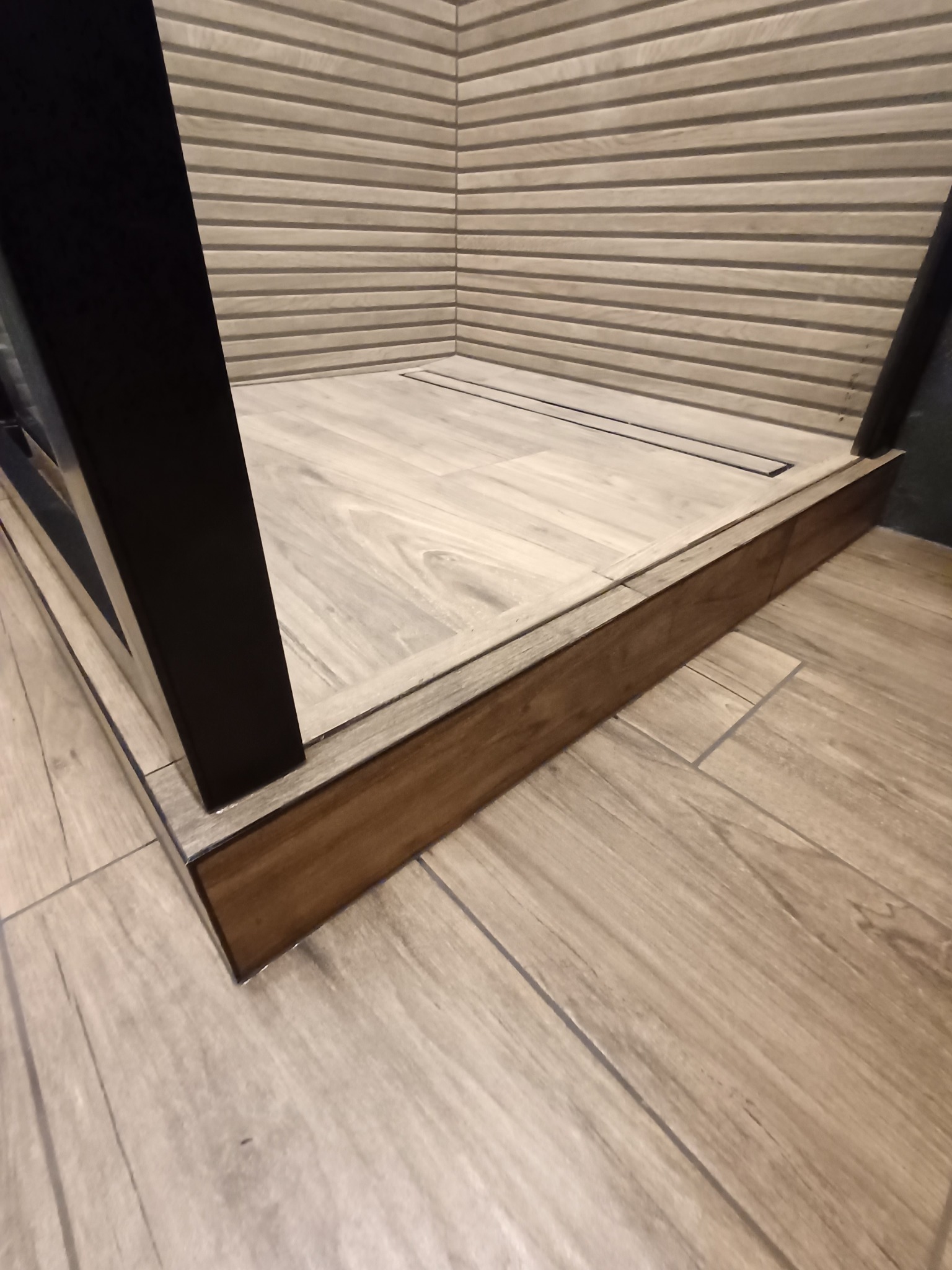 Nowoczesna kabina prysznicowa walk-in z drewnopodobnymi płytkami na ścianach i podłodze, z odpływem liniowym. Minimalistyczny design, ciepła kolorystyka, widok z dołu.