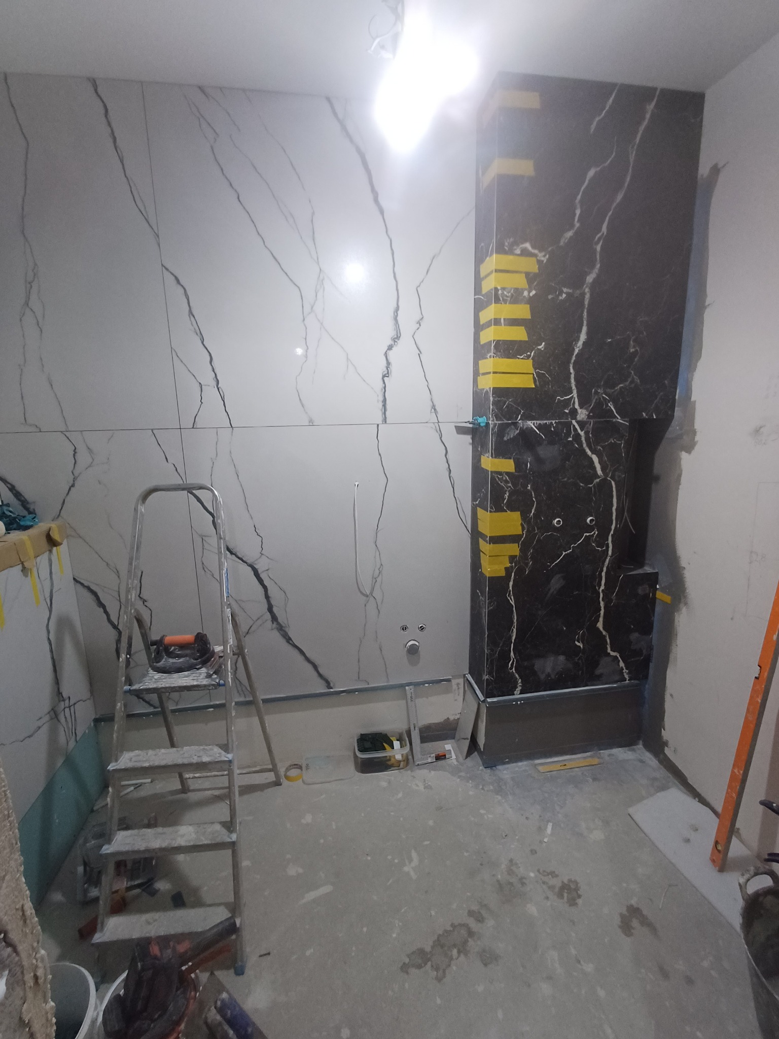 Łazienka w trakcie prac: białe i czarne płytki imitujące marmur na ścianach, drabina, poziomica. Żółte taśmy zabezpieczające na czarnych płytkach.