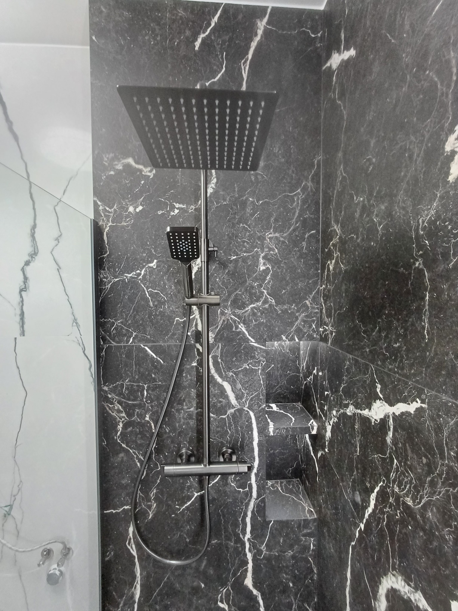 Nowoczesna kabina prysznicowa z deszczownicą i baterią w kolorze grafitowym, wykończona czarnym marmurem z białymi żyłami i półkami wnękowymi.