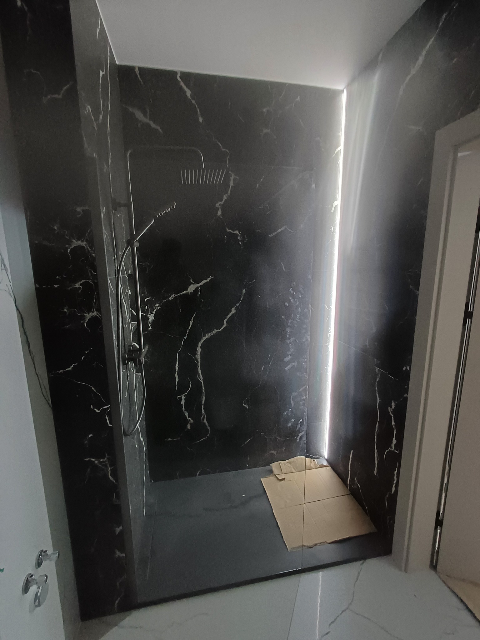 Kabina prysznicowa z czarnymi płytkami imitującymi marmur, wykończona w Ozorkowie. Widoczna deszczownica i szklana ścianka. Na podłodze karton ochronny.