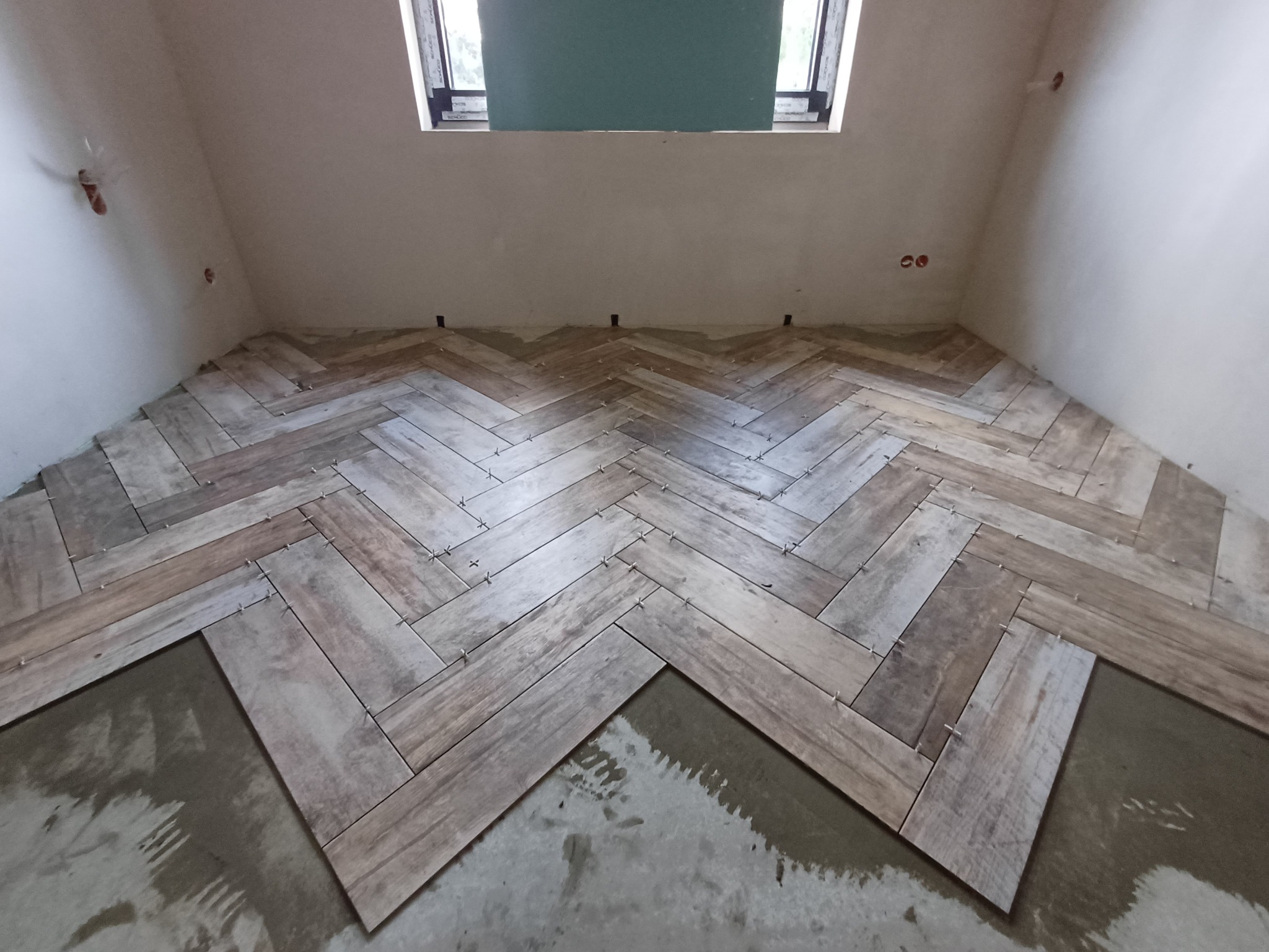 Układanie drewnopodobnych płytek w jodełkę na podłodze w pomieszczeniu z oknem dachowym. Widoczne krzyżyki dystansowe i fragmenty niepokrytej podłogi.