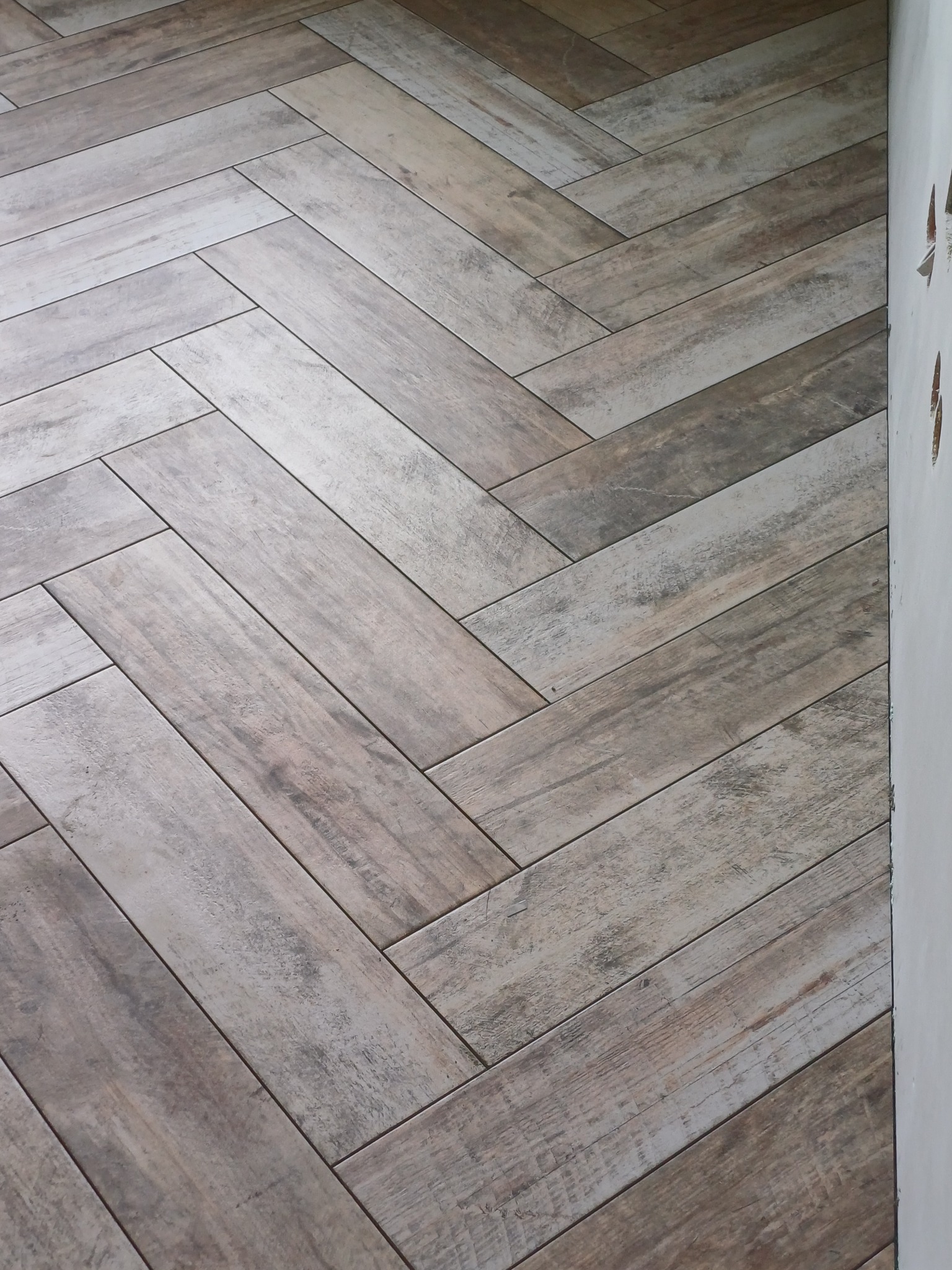 Ułożona w jodełkę podłoga z płytek imitujących drewno w odcieniach szarości i beżu, widoczna przy białej ścianie. Detal wykończenia wnętrza.