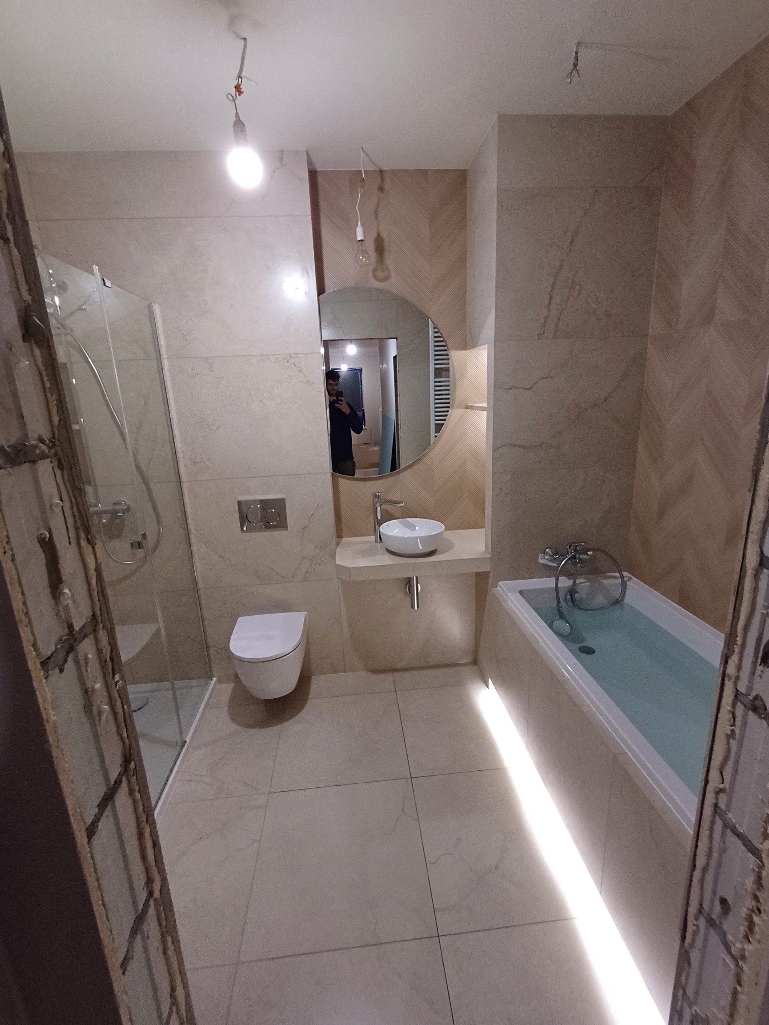 Jasna łazienka po remoncie z kabiną prysznicową, toaletą, okrągłym lustrem i wanną z podświetleniem LED. Ściany wyłożone płytkami w odcieniach beżu z wzorem jodełki.