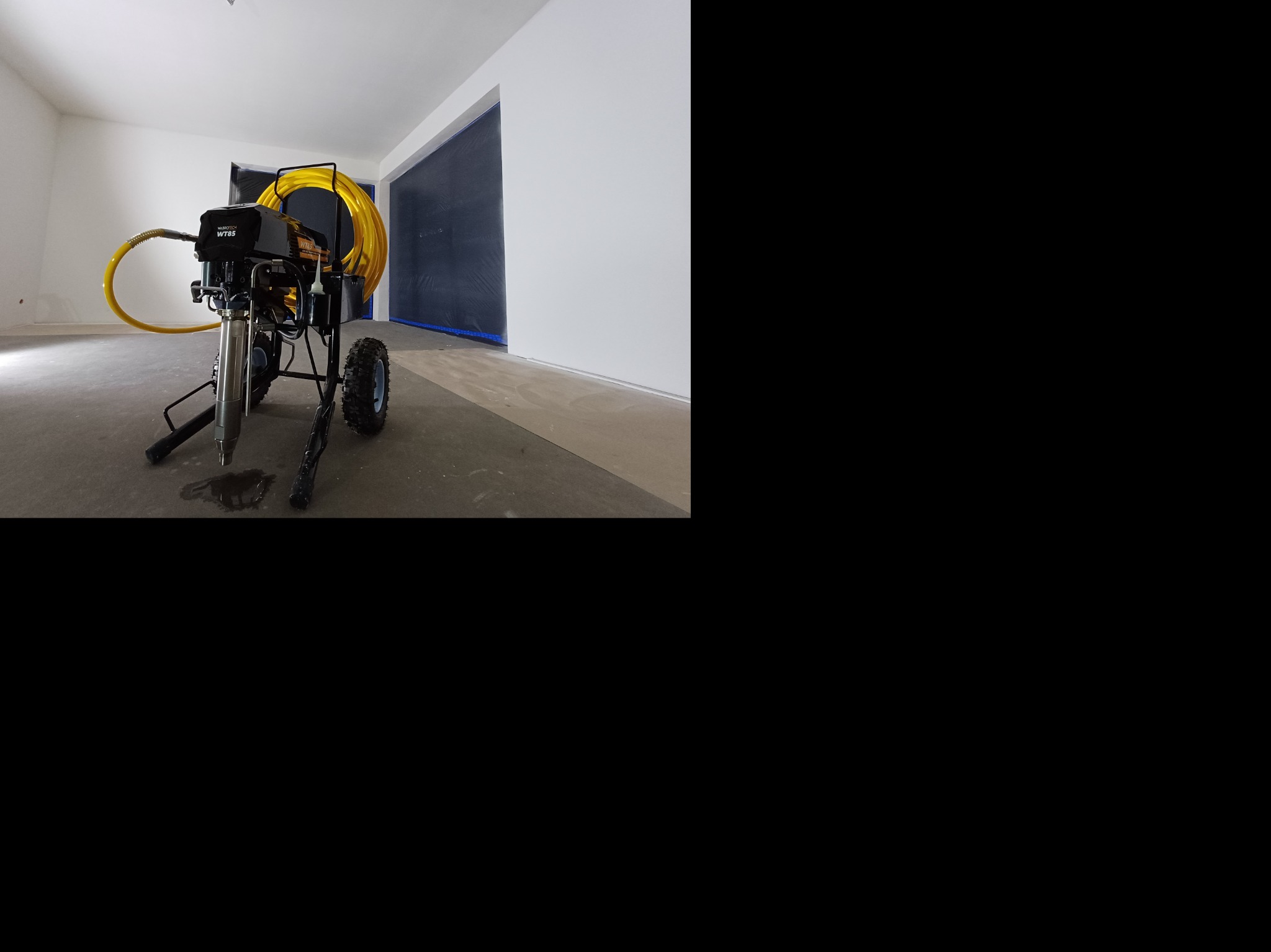 Agregat malarski z żółtym wężem w pustym, przygotowanym do malowania pomieszczeniu. Ściany białe, podłoga szara, okna zabezpieczone folią.