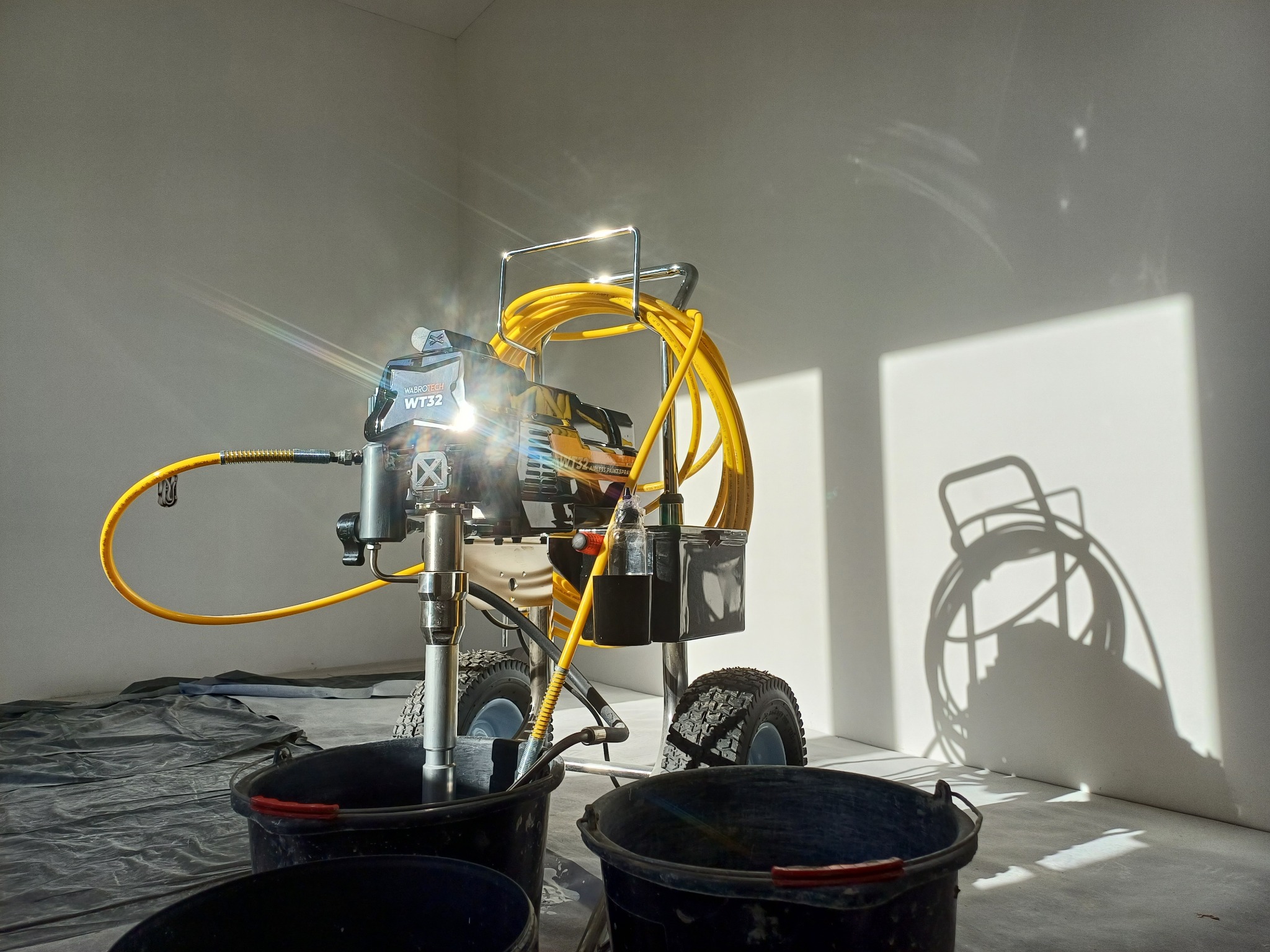 Profesjonalny agregat malarski Wabrotech WT32 z żółtym wężem i trzema wiadrami na folii malarskiej, oświetlony promieniami słońca w pustym pomieszczeniu.