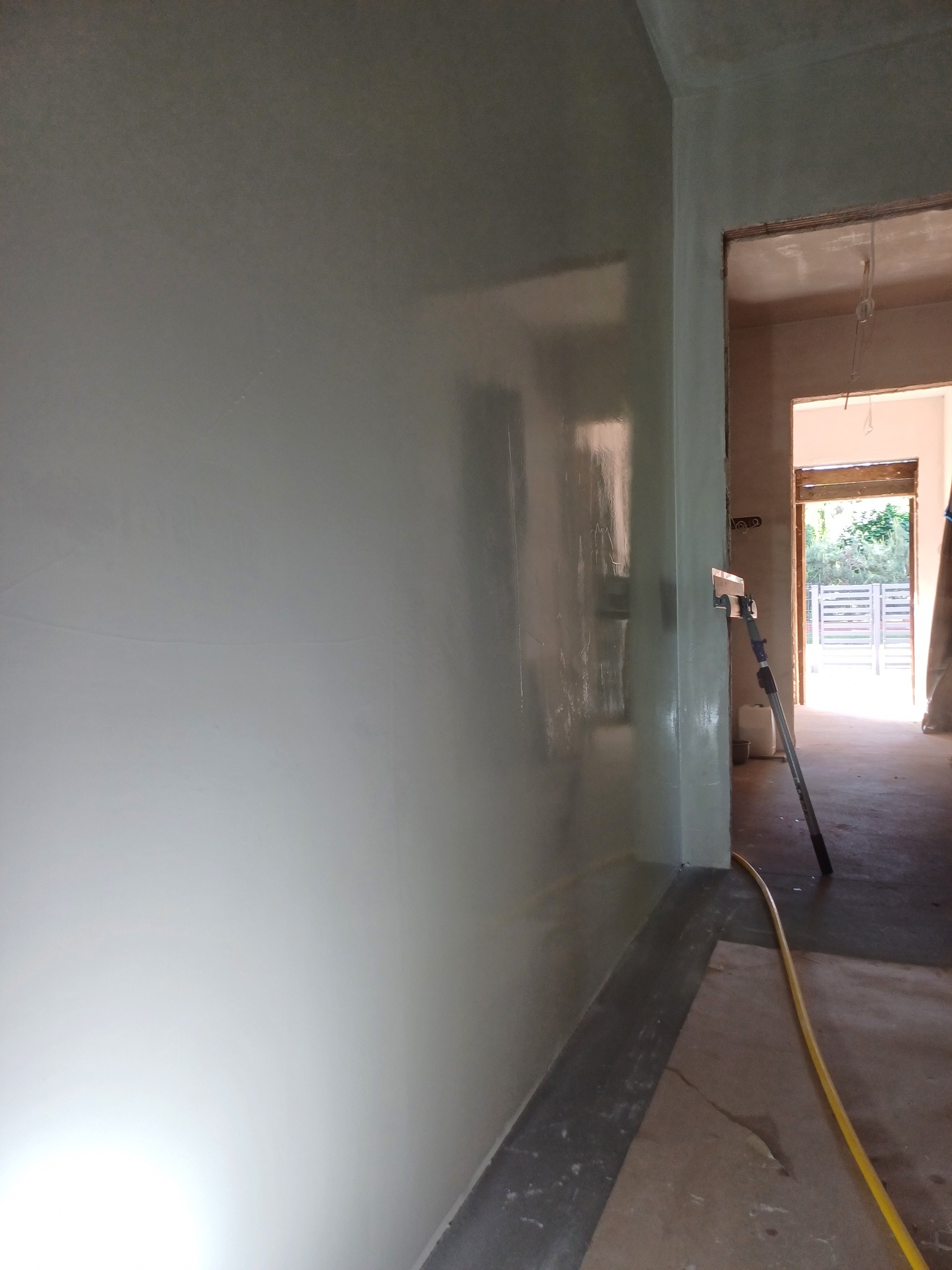 Świeżo nałożona, mokra gładź na ścianie w trakcie prac wykończeniowych. Widoczny sprzęt do rozprowadzania gładzi i fragment węża. W tle widoczne drzwi i zieleń.