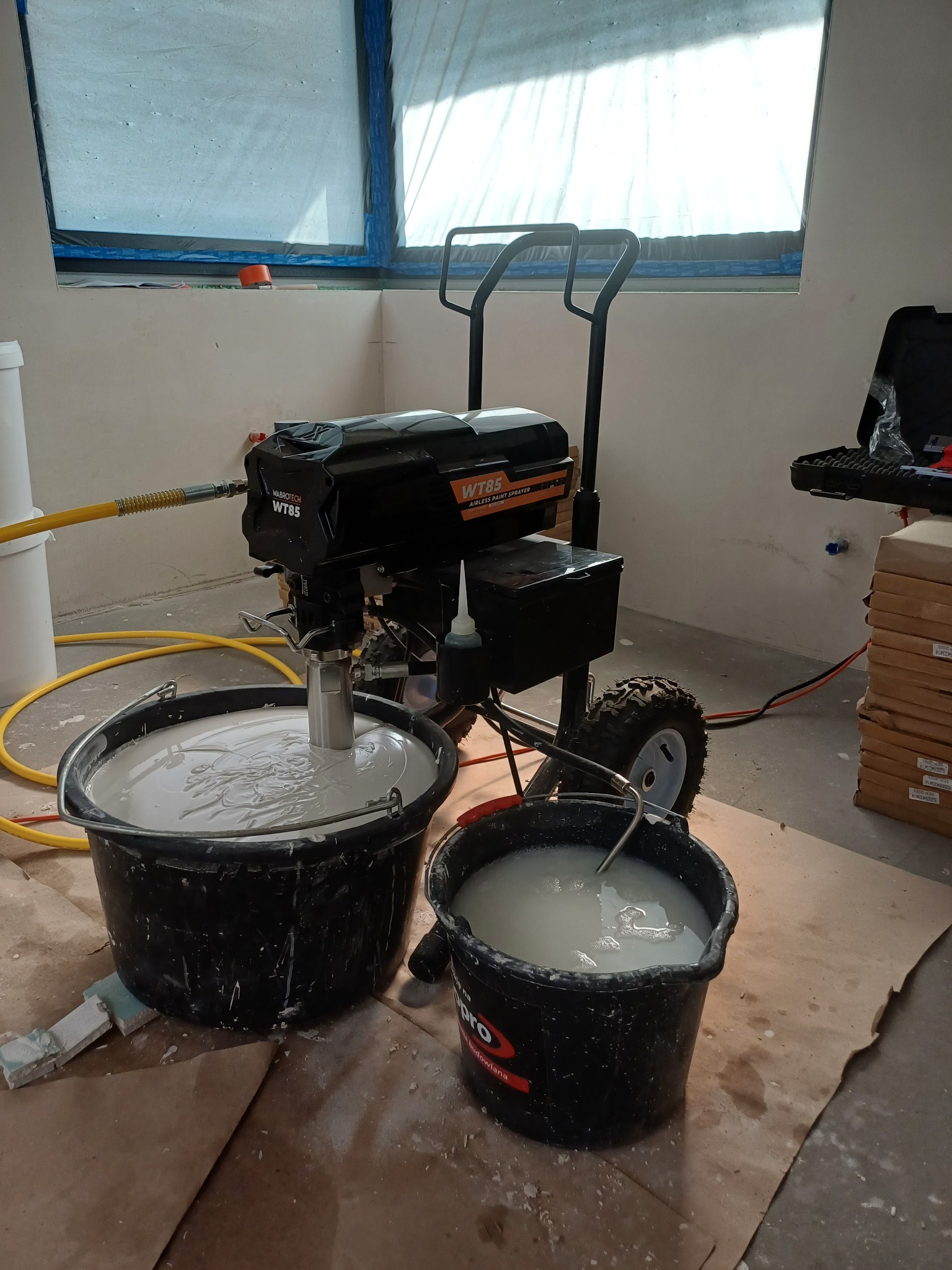 Agregat malarski WABROTECH WT85 z dwoma wiadrami z farbą/gładzią, przygotowany do pracy w pomieszczeniu z oknem zabezpieczonym folią malarską. Widoczne kartony z płytami.