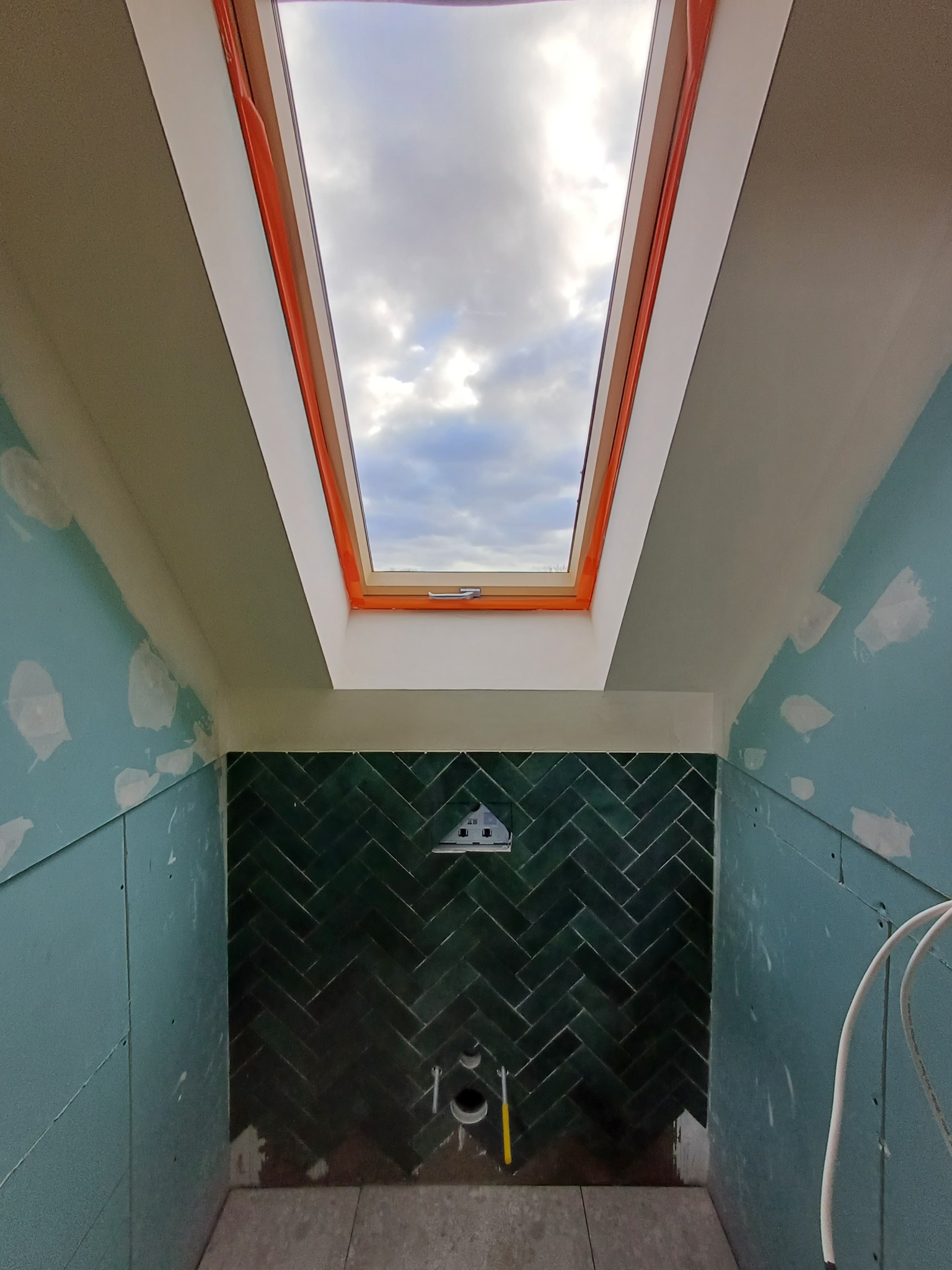 Łazienka w trakcie remontu: zielona glazura ułożona w jodełkę na ścianie, sufit z oknem dachowym, ściany wyłożone płytami gipsowo-kartonowymi, widoczne instalacje.