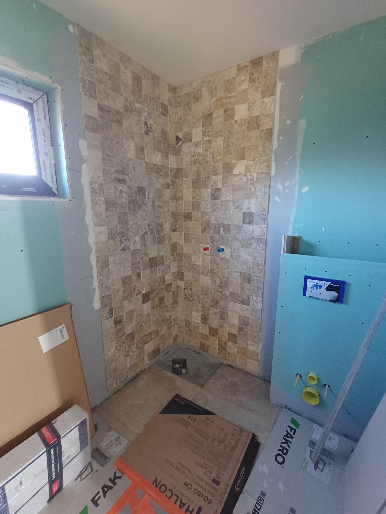 Fragment łazienki w trakcie wykańczania: ułożone płytki mozaikowe na ścianie prysznica, obok ściana z płyty gipsowej z instalacją podtynkową WC. Widoczne kartony z płytkami.