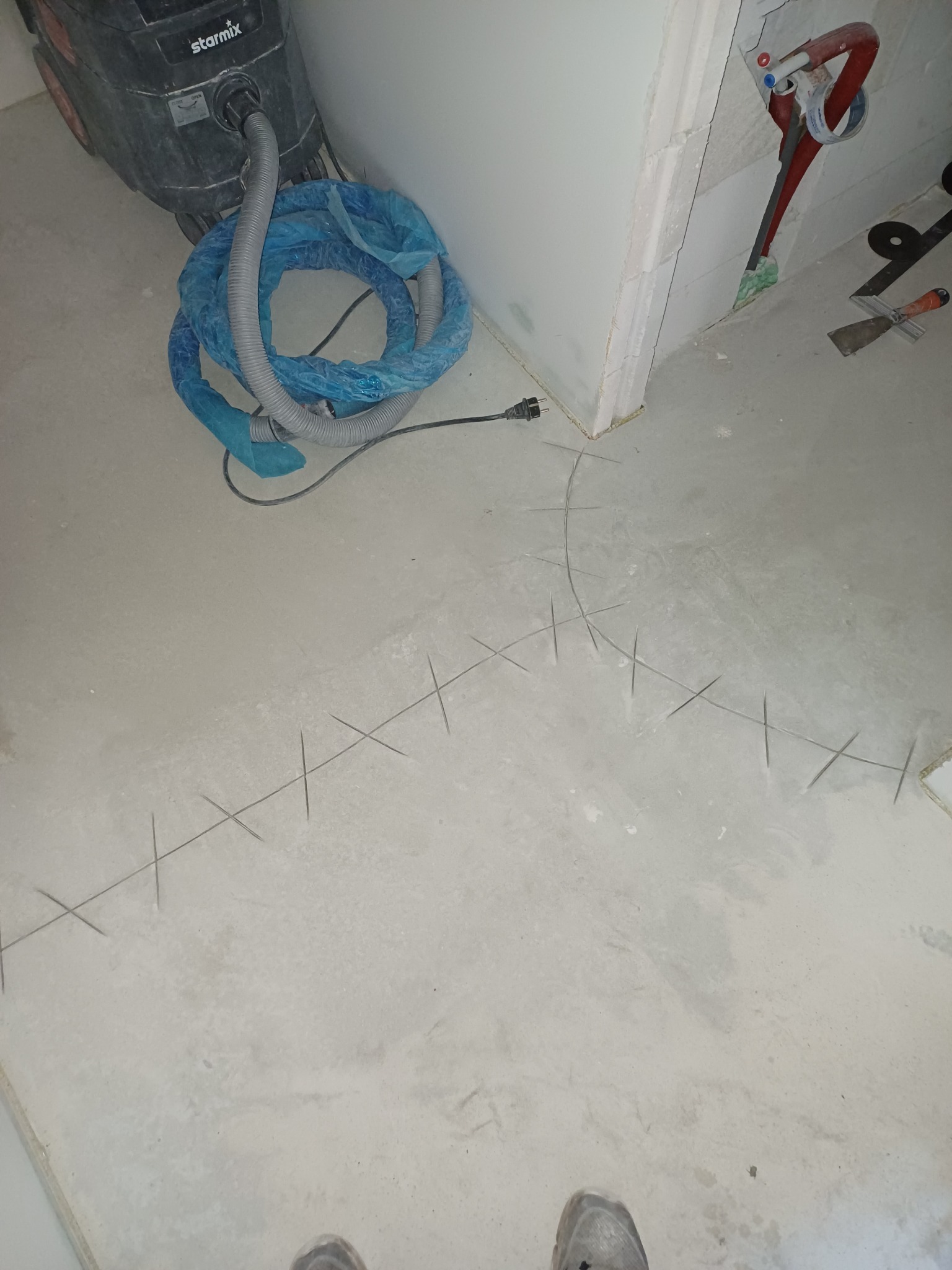 Przygotowanie podłoża pod wylewkę: zaznaczone krzyżykami punkty poziomowania, odkurzacz przemysłowy i narzędzia glazurnicze w tle. Wnętrze w trakcie prac.