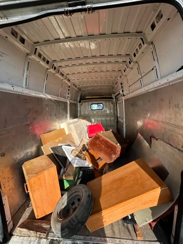 Wnętrze furgonetki wypełnionej różnorodnymi przedmiotami, w tym meblami, oponą i innymi elementami. Widok z tyłu pojazdu, ukazujący załadunek.