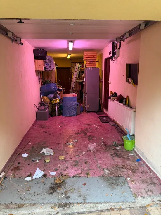 Zaniedbane pomieszczenie gospodarcze z różową poświatą, zagracone przedmiotami i śmieciami na podłodze z płytek. Widoczne regały, sprzęt AGD i osobiste rzeczy.